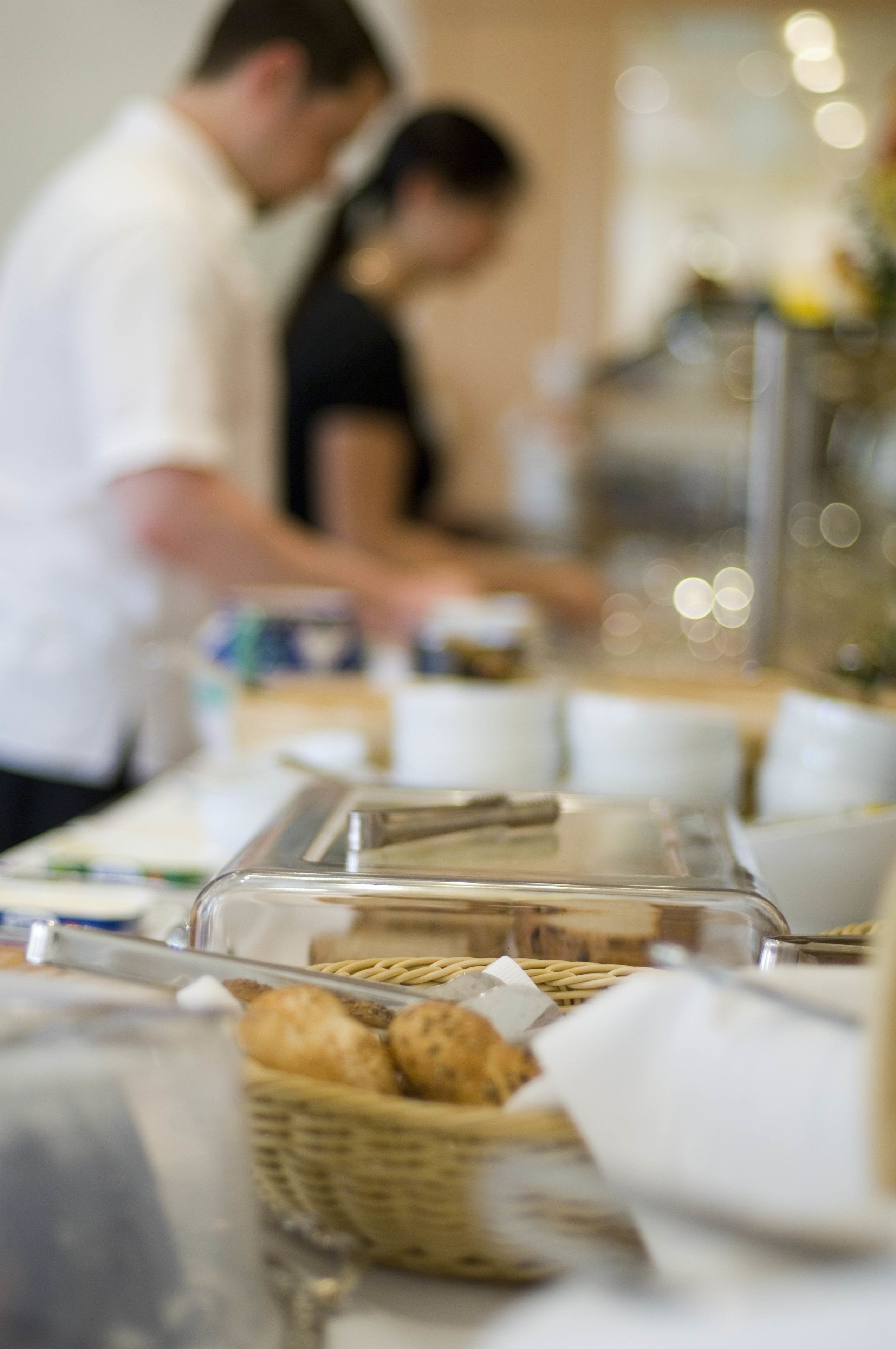 A buffet setup with a clear glass container holding bread rolls, a basket of bread rolls, and a white napkin.