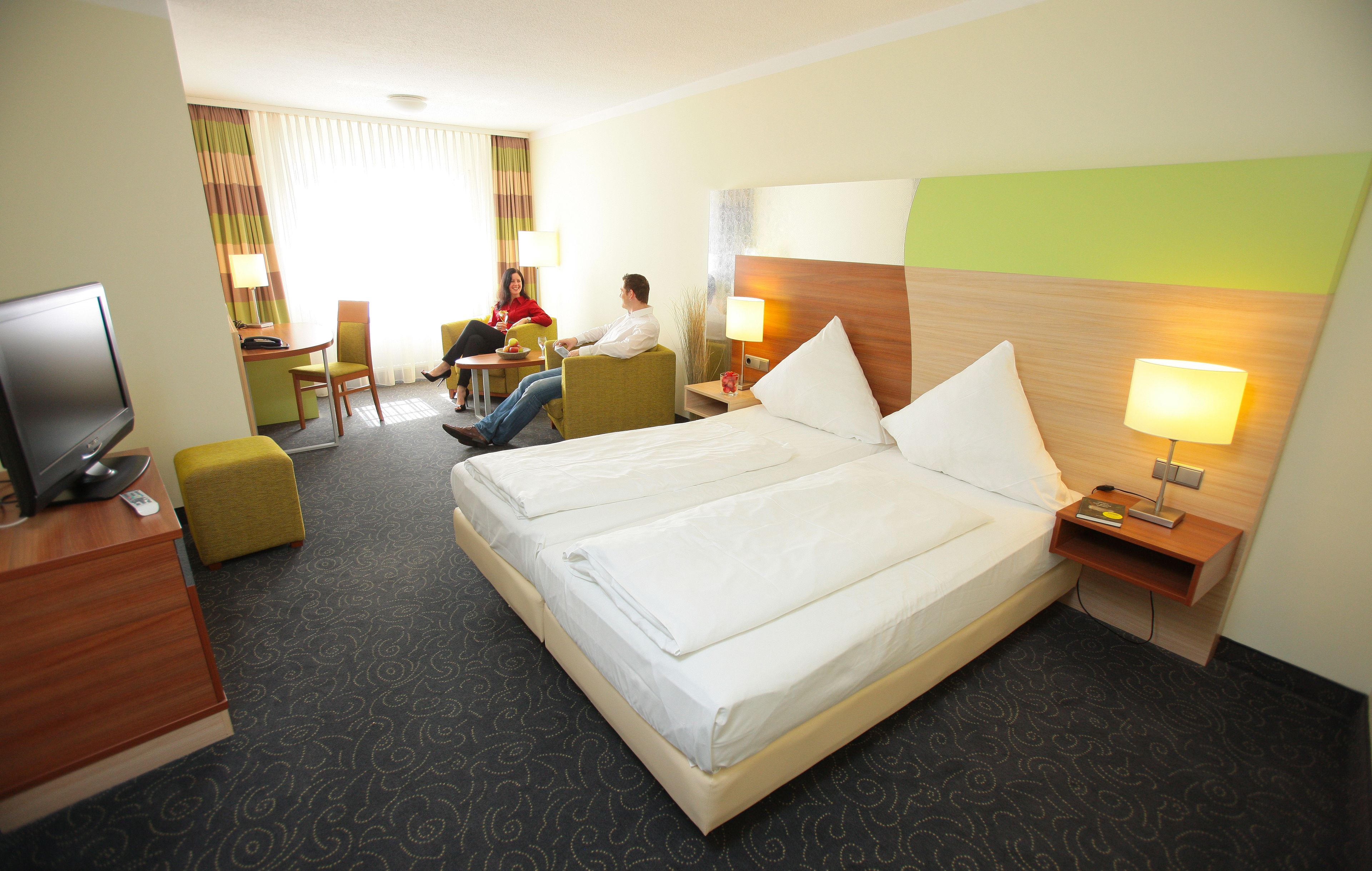 Habitación de hotel con dos camas, un escritorio, televisor y una zona de estar con una persona.