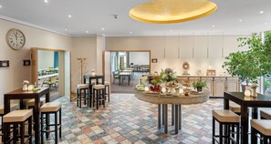 Un hall d’hôtel moderne avec une table ronde centrale où sont disposés des plats et des boissons, des tabourets de bar et une grande horloge au mur.
