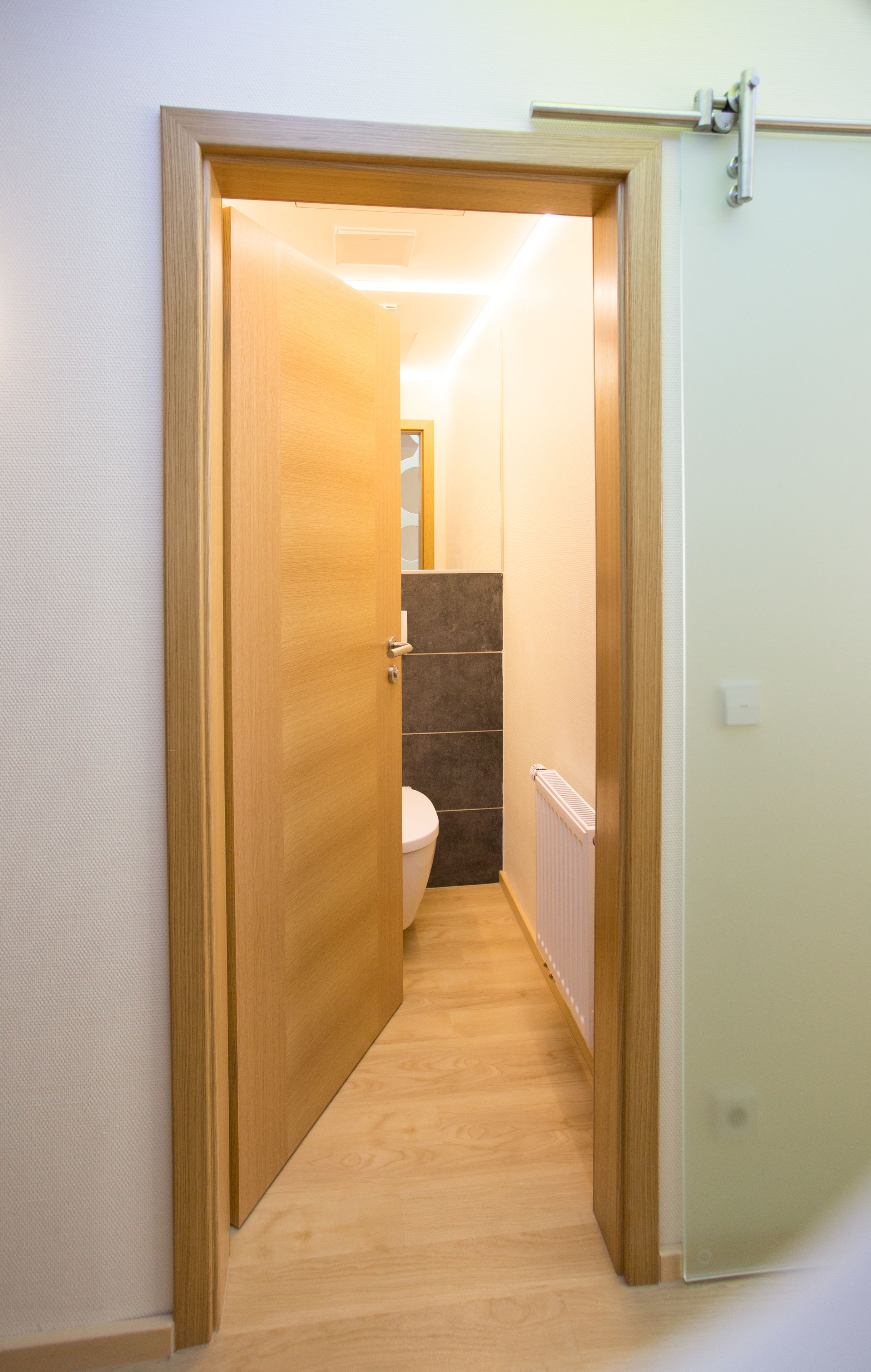 Ein schmaler Flur mit einer Holztür, die zu einem Badezimmer führt. Das Badezimmer ist mit Toilette und Heizkörper ausgestattet.