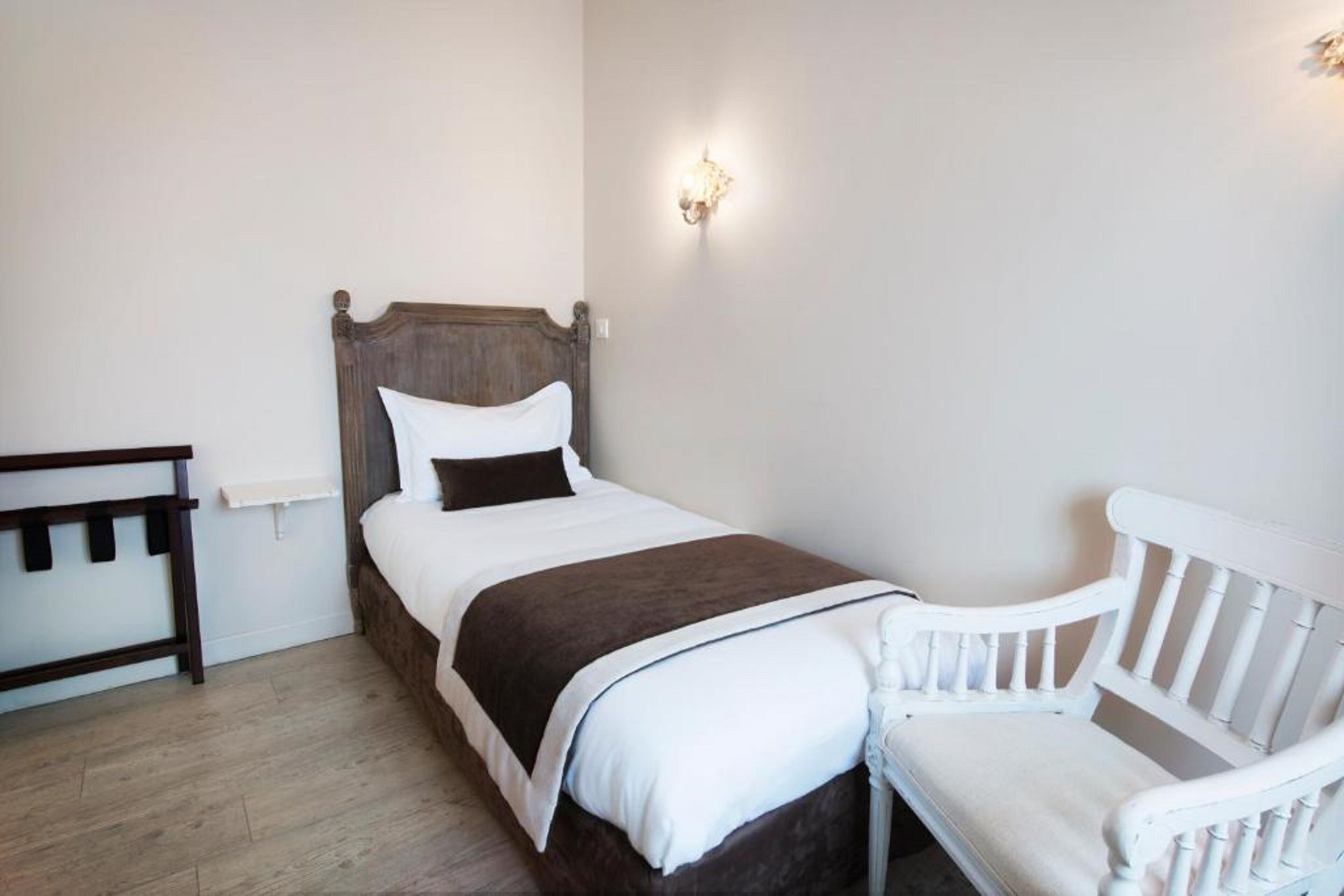 A bedroom with a wooden bed, white bedding, a white chair, and a wall-mounted light fixture.