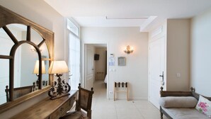 A hallway with a wooden bench, a desk with a lamp, and a large mirror. There is a door on the right and a wall-mounted light fixture above it. - Hôtel Le Mas Saint-Joseph (St.-Rémy-de-Provence)