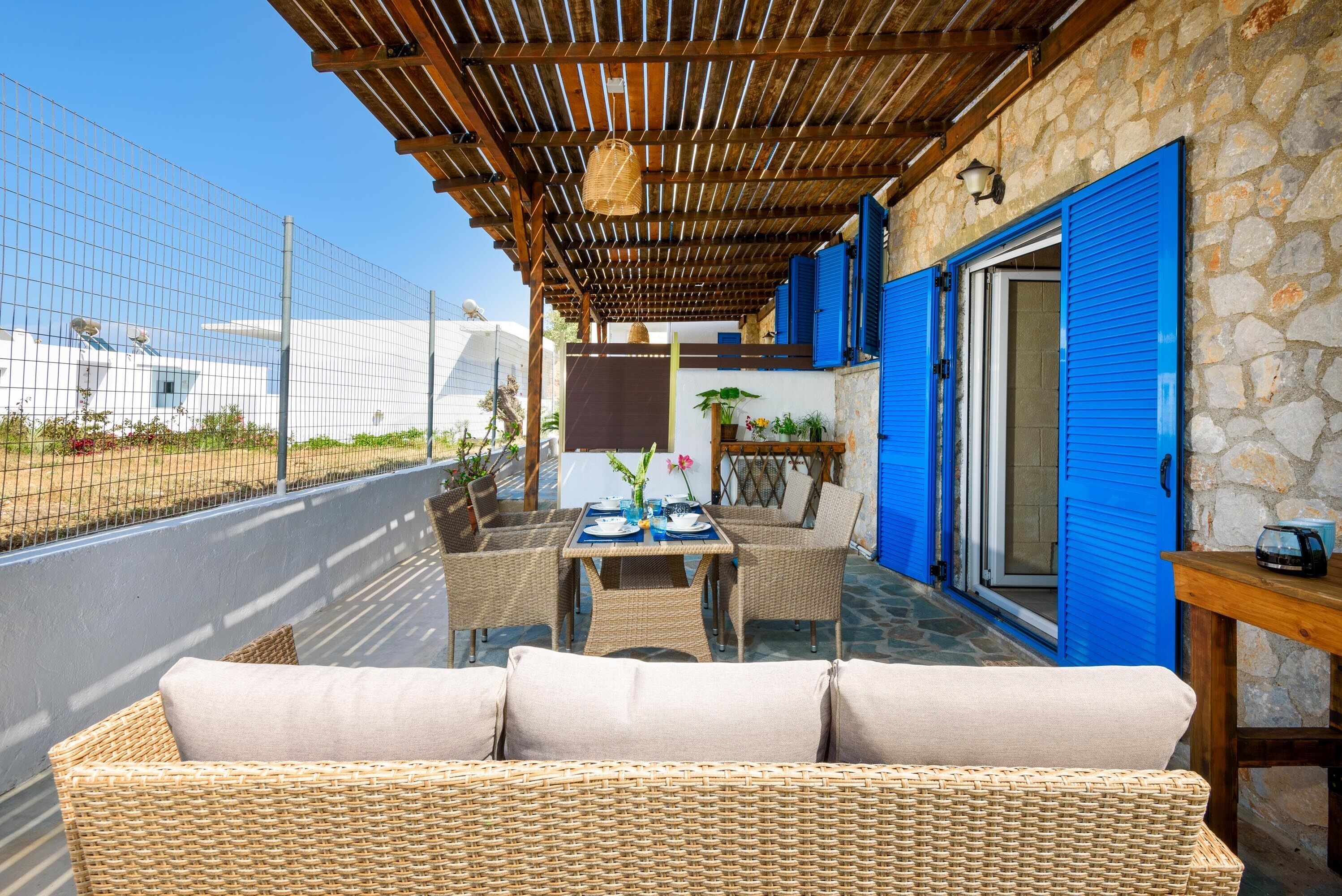 Eine Terrasse im Freien mit Korbmöbeln, einem Esstisch und einer Steinmauer mit blauen Fensterläden.