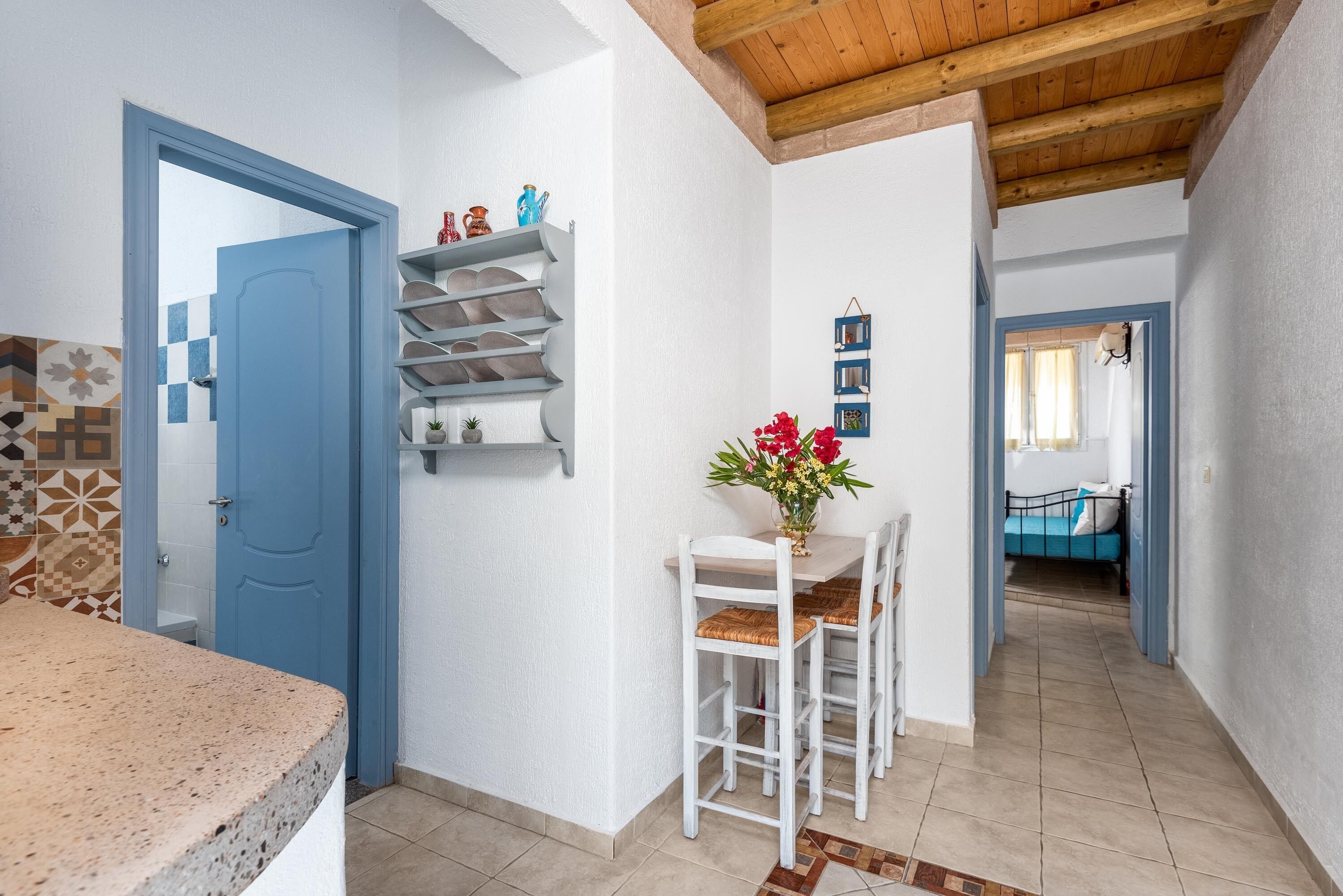 A modern kitchen with a blue door, a dining area with white chairs and a table, and a living area with a blue sofa.