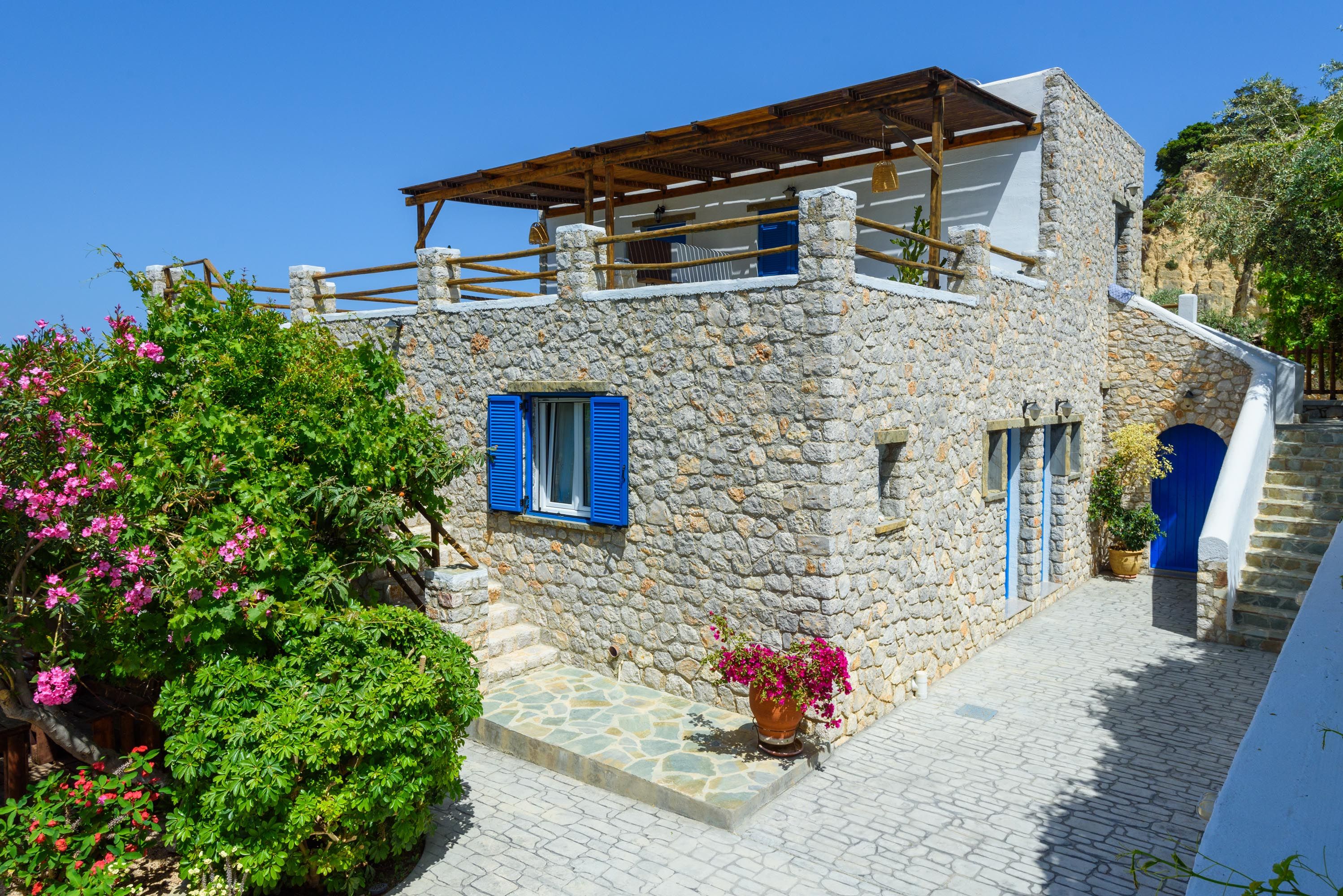 A stone building with a wooden balcony, a window with blue shutters, and a potted plant with pink flowers.
