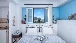 Un dormitorio con cama, ventana con cortinas y vista al mar, además de una mesita de noche pequeña.