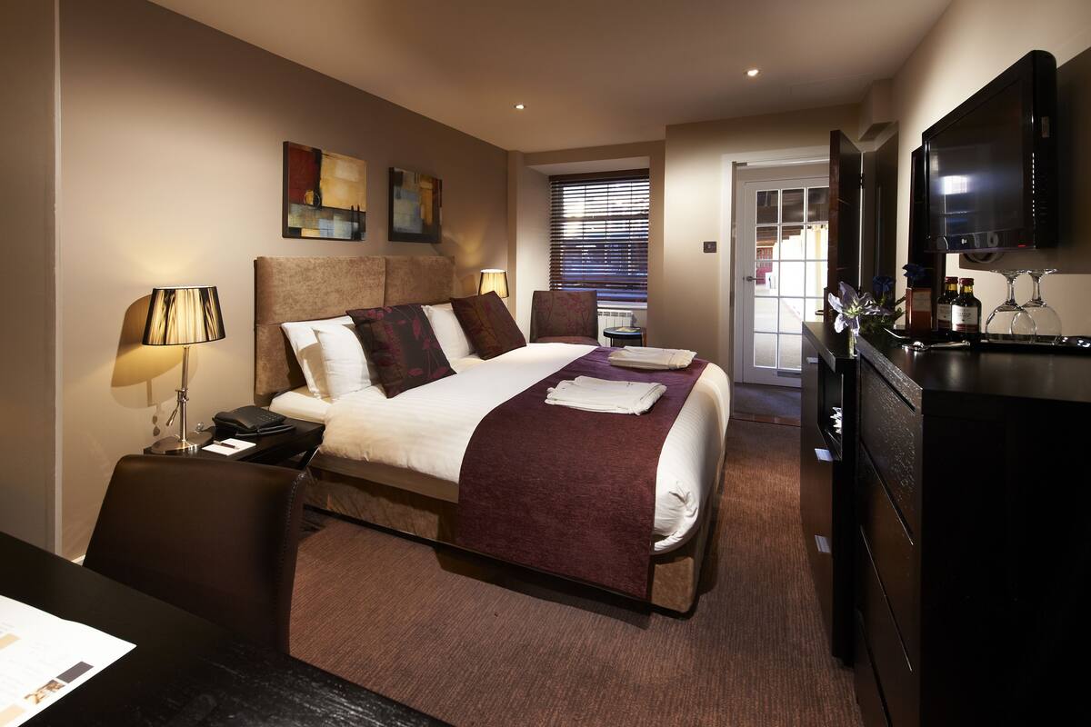 A hotel room with a bed, bedside tables, a desk, a TV, and a window with blinds.