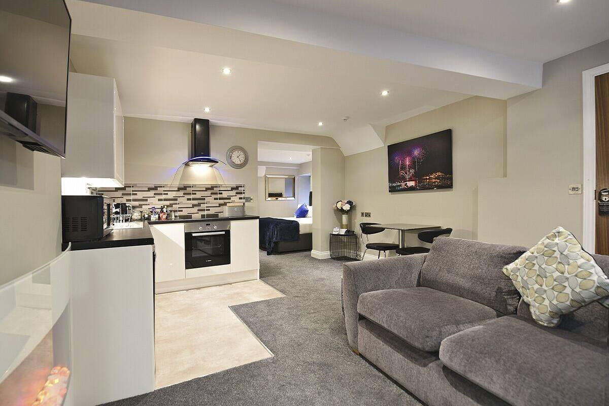 A modern living room with a grey sofa, a small dining table, and a kitchen area with a built-in oven and microwave.