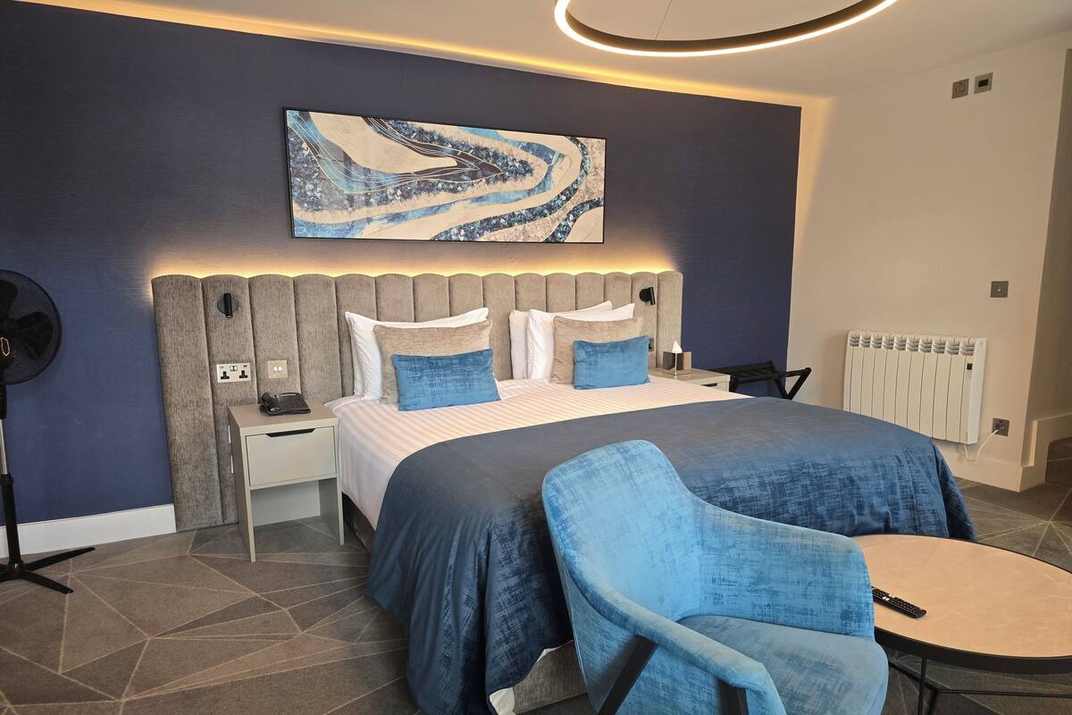 A modern bedroom with a large bed, a headboard with built-in lighting, a bedside table with a lamp, and a chair with a table.