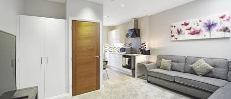 A modern living room with a grey sofa, a wooden door, and a kitchen in the background.