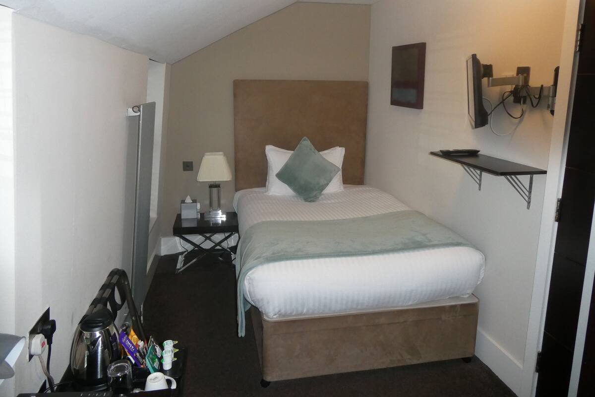 A small, neatly arranged hotel room with a single bed, bedside table, and a TV mounted on the wall.