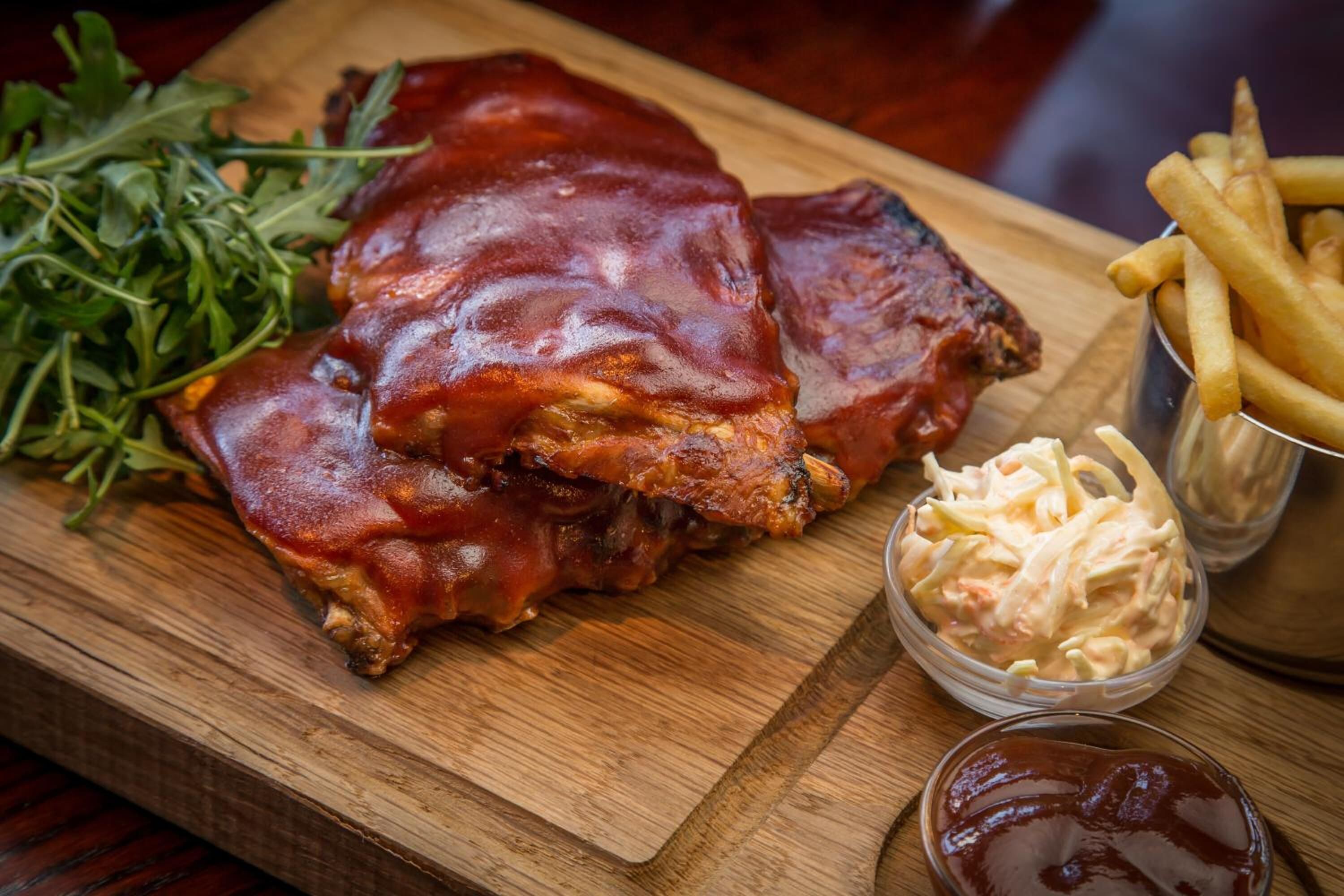 Una tabla de madera con costillas a la parrilla, papas fritas, ensalada de col y un pequeño recipiente de kétchup.