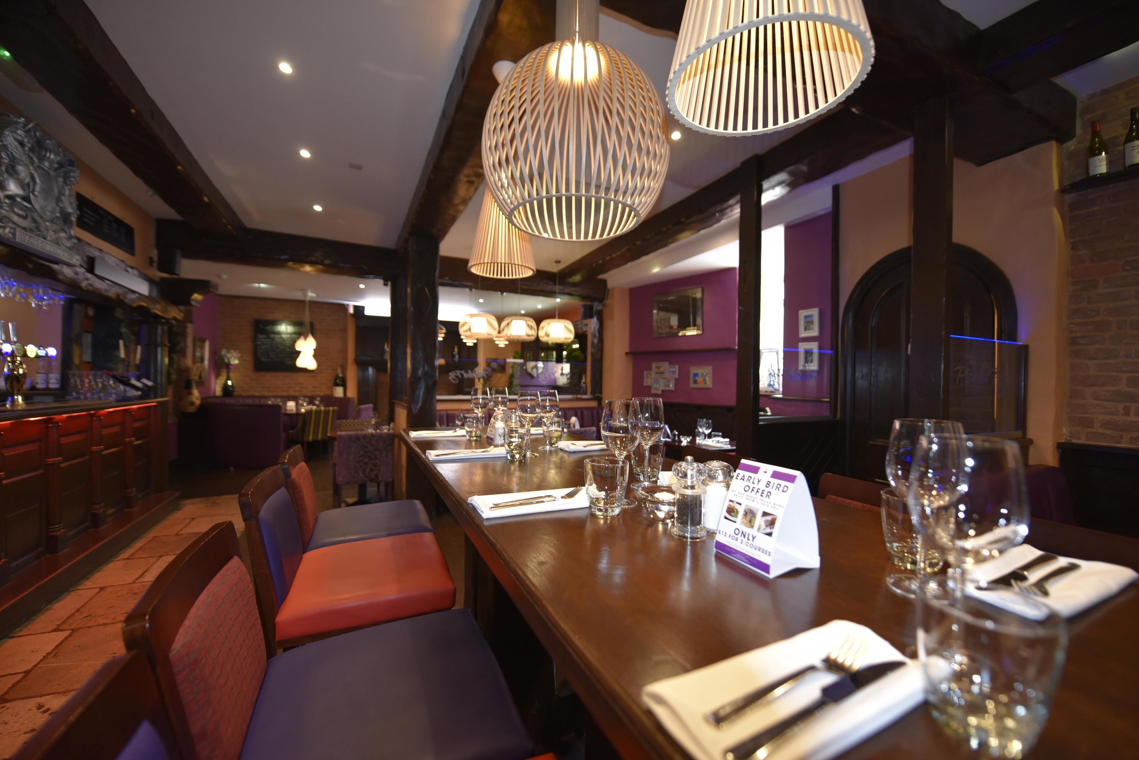 Un restaurante con mesas de madera preparadas para comer, una zona de bar y una pared morada con cuadros enmarcados.