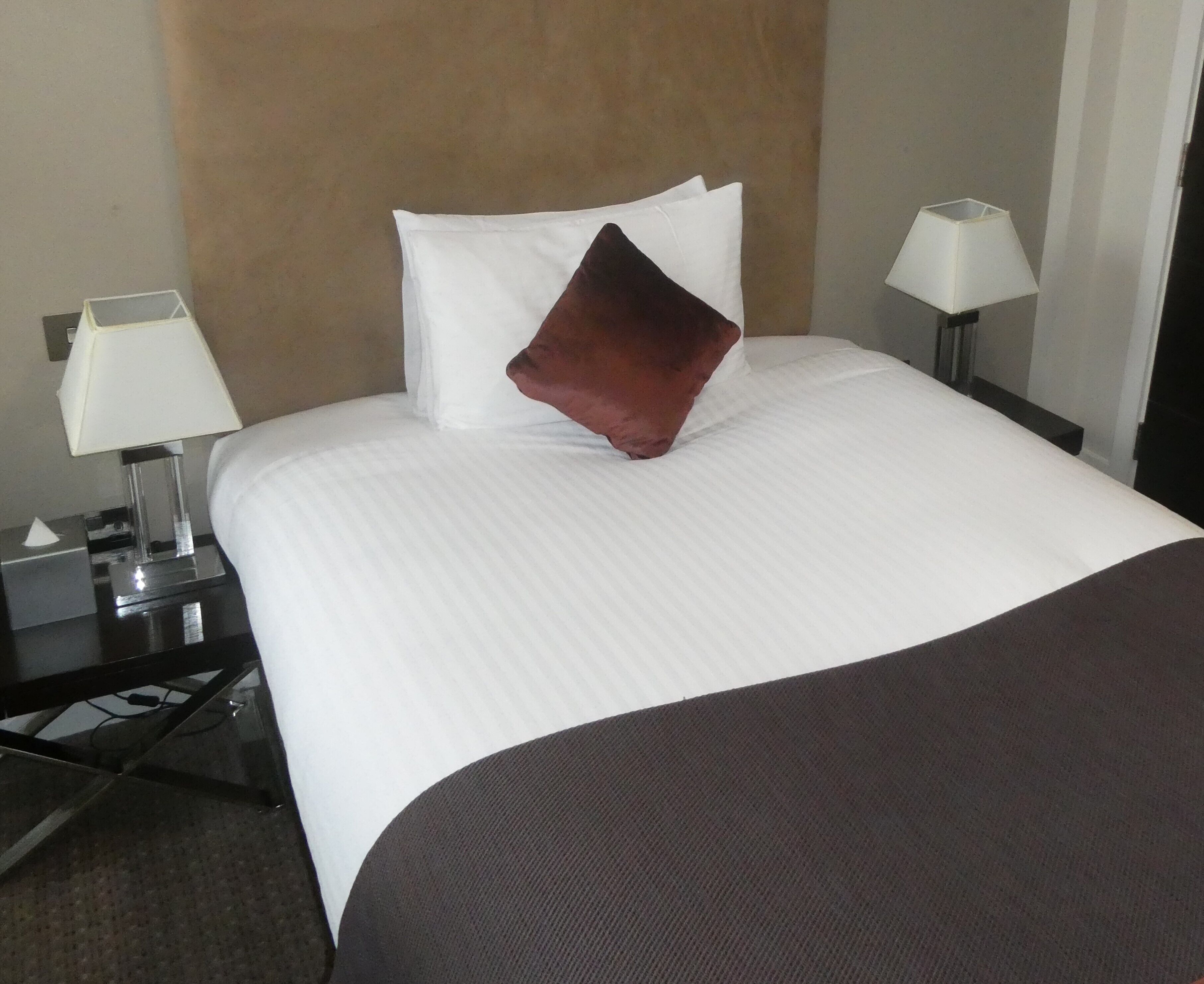 Un lit bien fait, avec du linge de lit blanc, une tête de lit marron et un seul oreiller marron. Il y a deux tables de chevet, chacune avec une lampe et une boîte de mouchoirs.