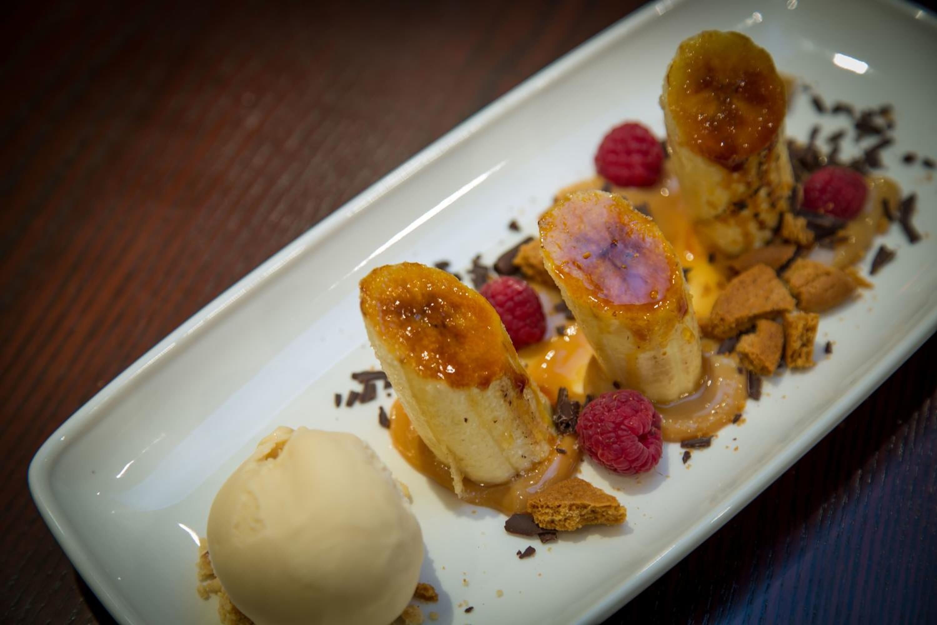 En desserttallrik med glass och karamelliserade bananer, serverad med hallon och hyvlad choklad.