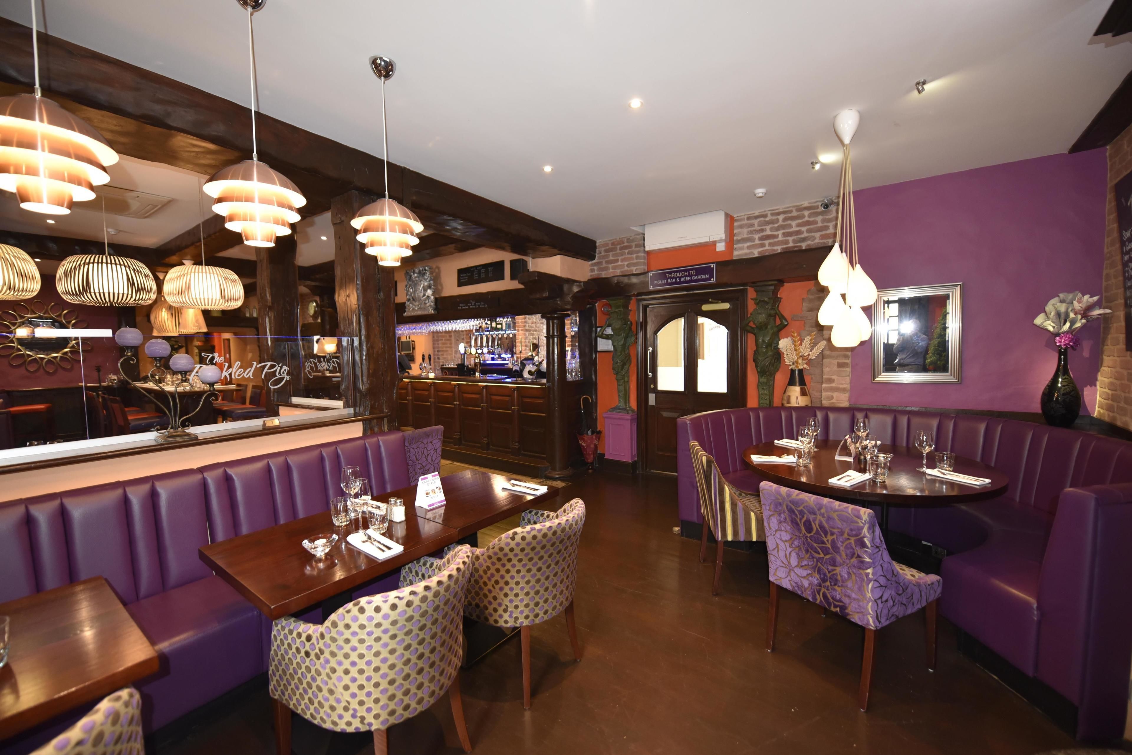 A restaurant with purple booths and tables, hanging pendant lights, and a brick wall.