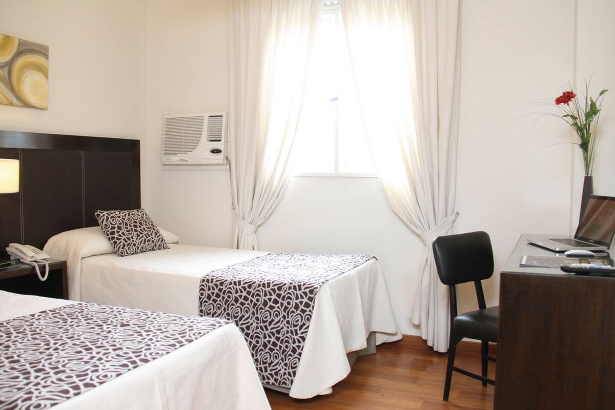 Une chambre d’hôtel avec deux lits, un bureau avec un ordinateur, une chaise, un téléphone et une fenêtre avec des rideaux.
