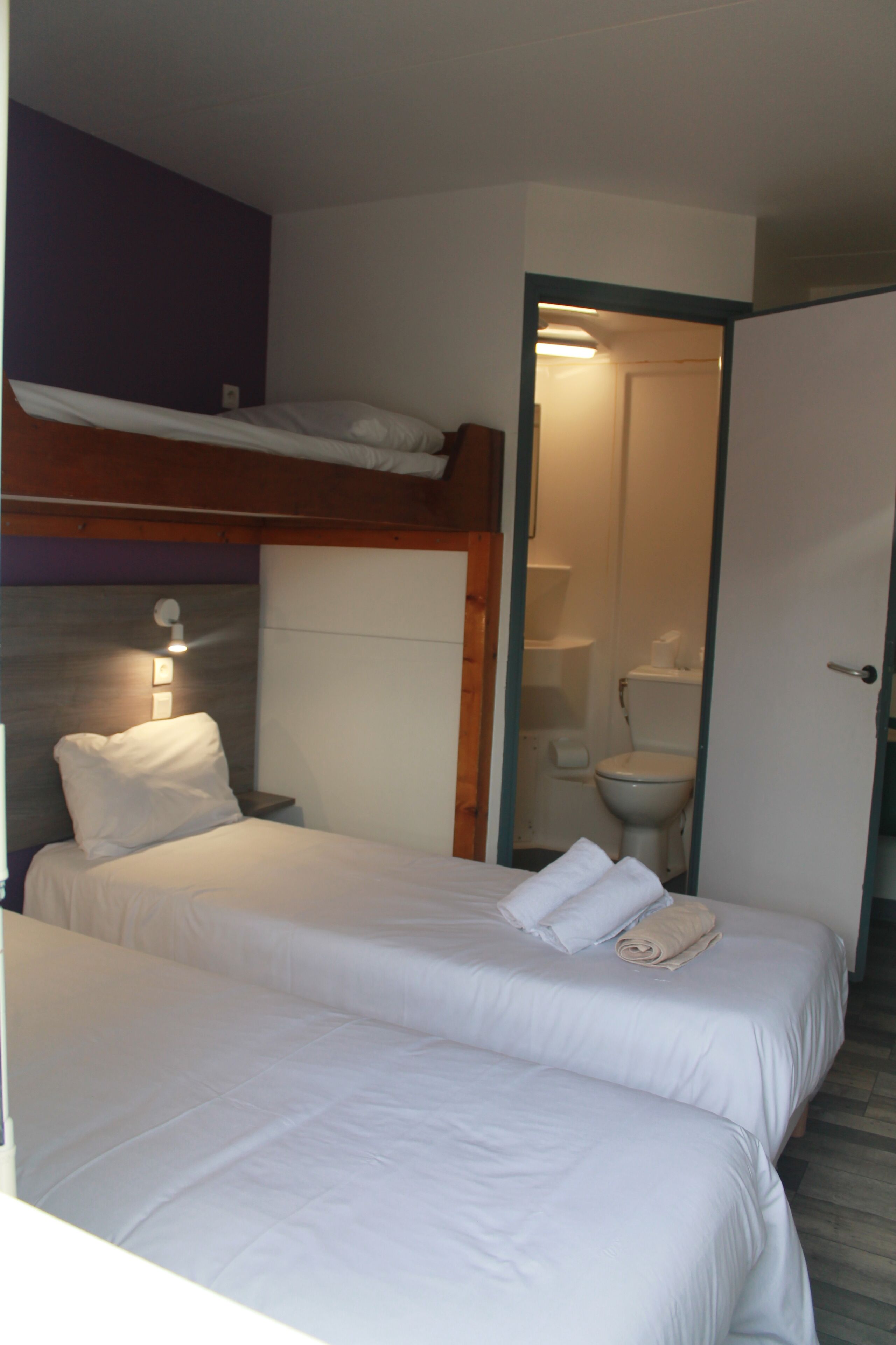 Een hotelkamer met twee bedden, een houten hoofdbord, een nachtkastje, een wandlamp en een badkamer die door een open deur zichtbaar is.