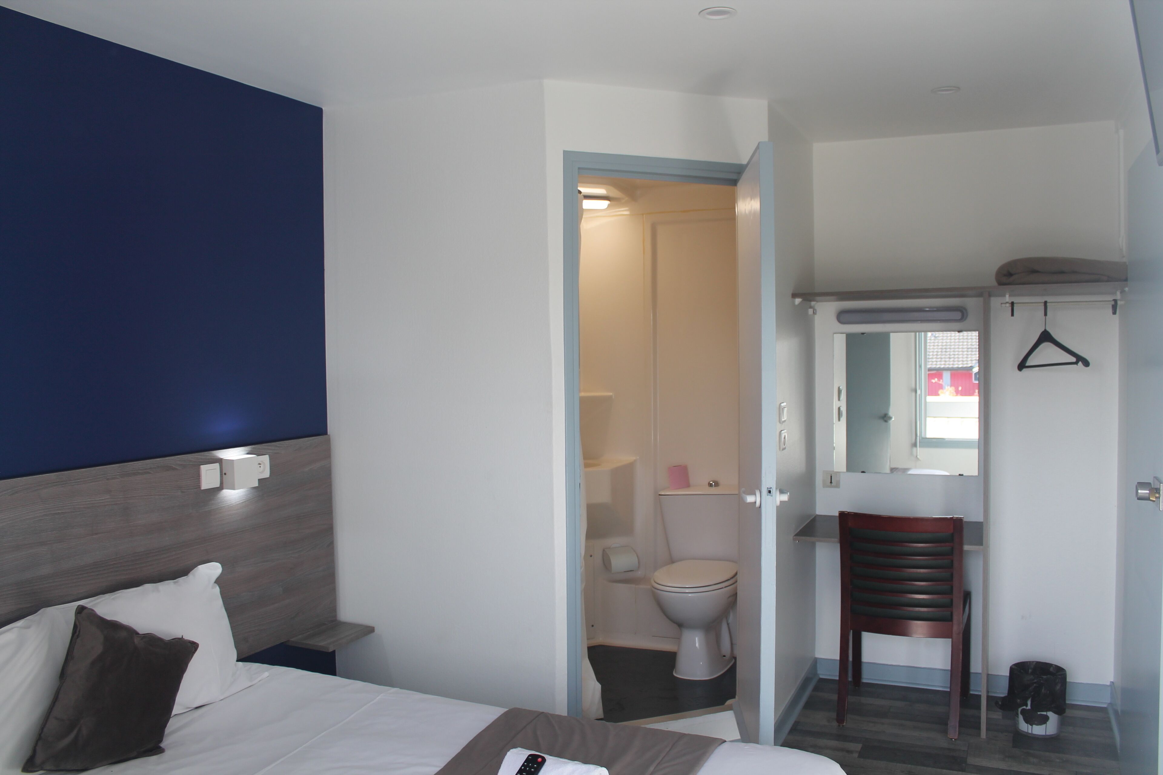 Une chambre d’hôtel avec un lit, une chaise, un bureau avec un miroir et une salle de bain visible à travers une porte ouverte.