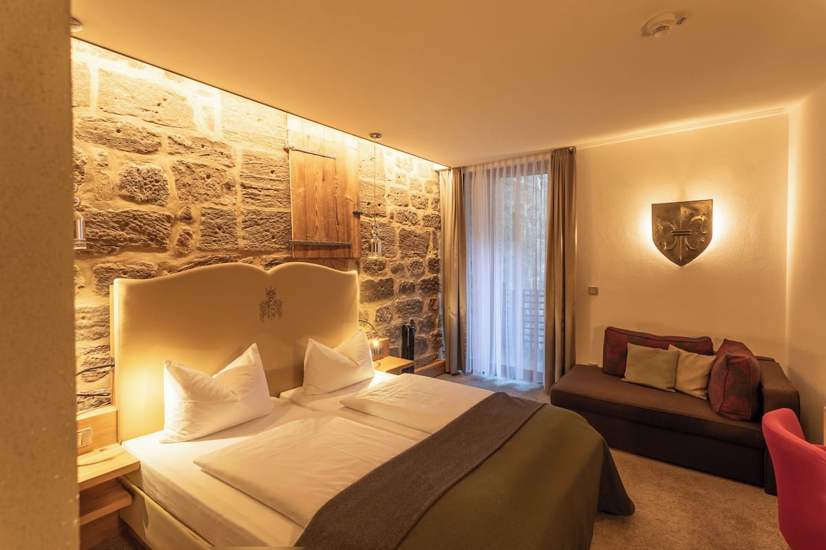 A hotel room with a stone wall, a bed with white linens, a wooden headboard, a nightstand, a red chair, and a wall-mounted shield-shaped light fixture.