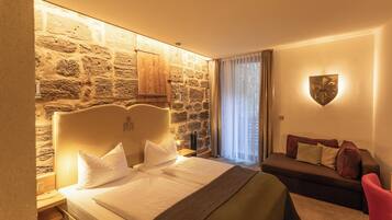 A hotel room with a stone wall, a bed with white linens, a wooden headboard, a nightstand, a red chair, and a wall-mounted shield-shaped light fixture.