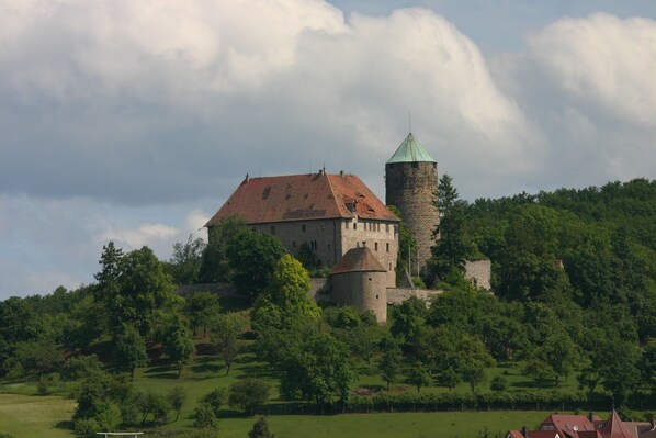 A historic castle with a prominent tower and a large building with a red roof, surrounded by lush greenery.