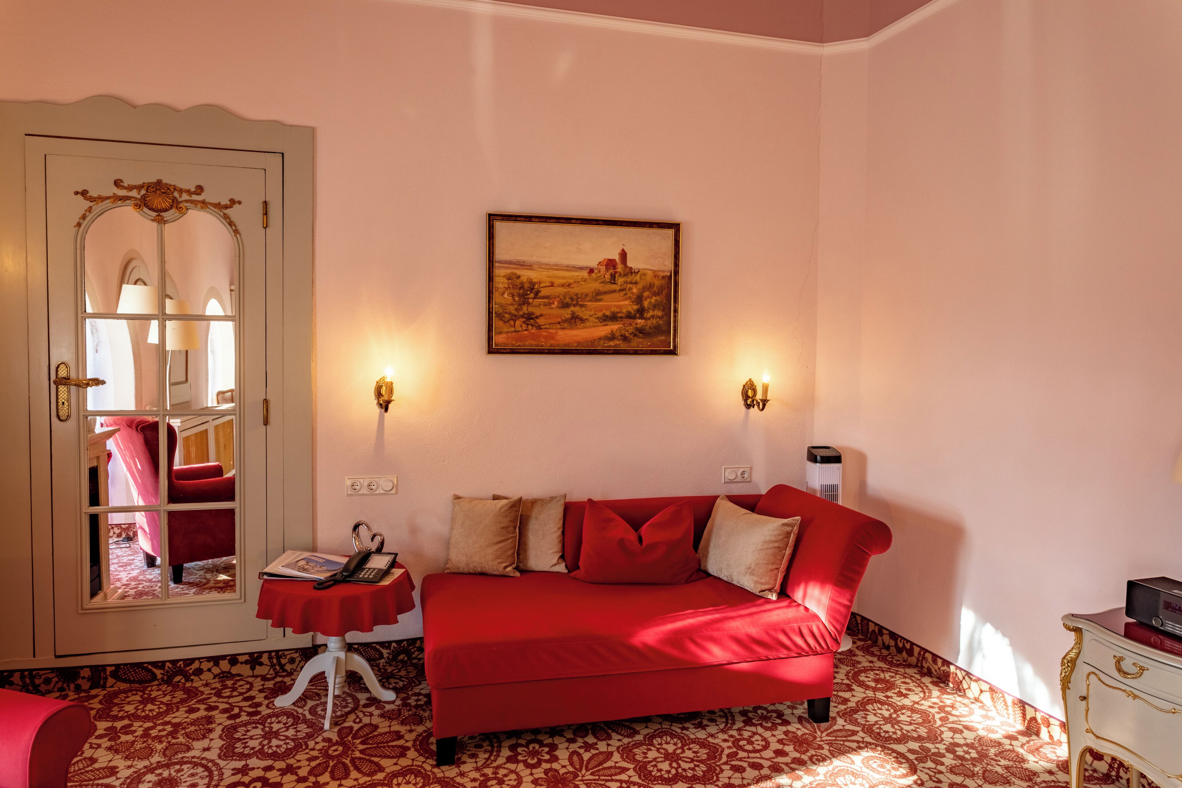 A room with a red sofa, a small table with a book and a phone, a painting on the wall, and a door with a mirror.