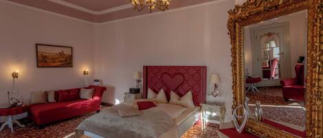 A bedroom with a large bed, a red sofa, a chandelier, and a mirror with an ornate frame.