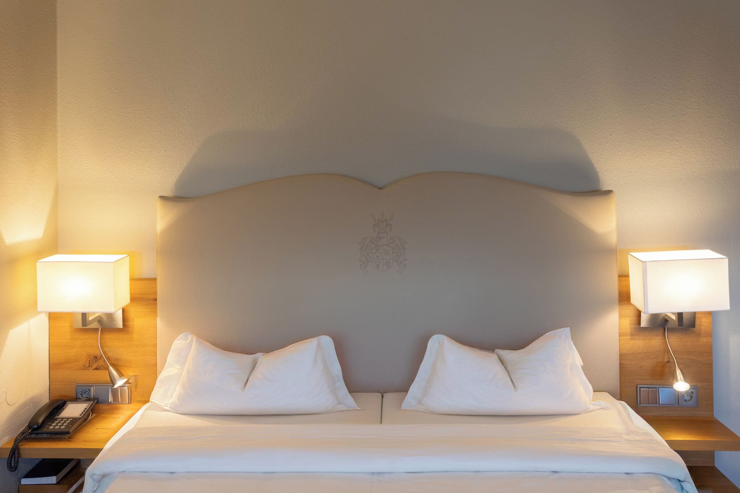 Ein Hotelzimmer mit einem großen Bett, zwei Nachttischlampen, einem Nachttisch mit Telefon und einem dekorativen Wappen am Kopfteil.