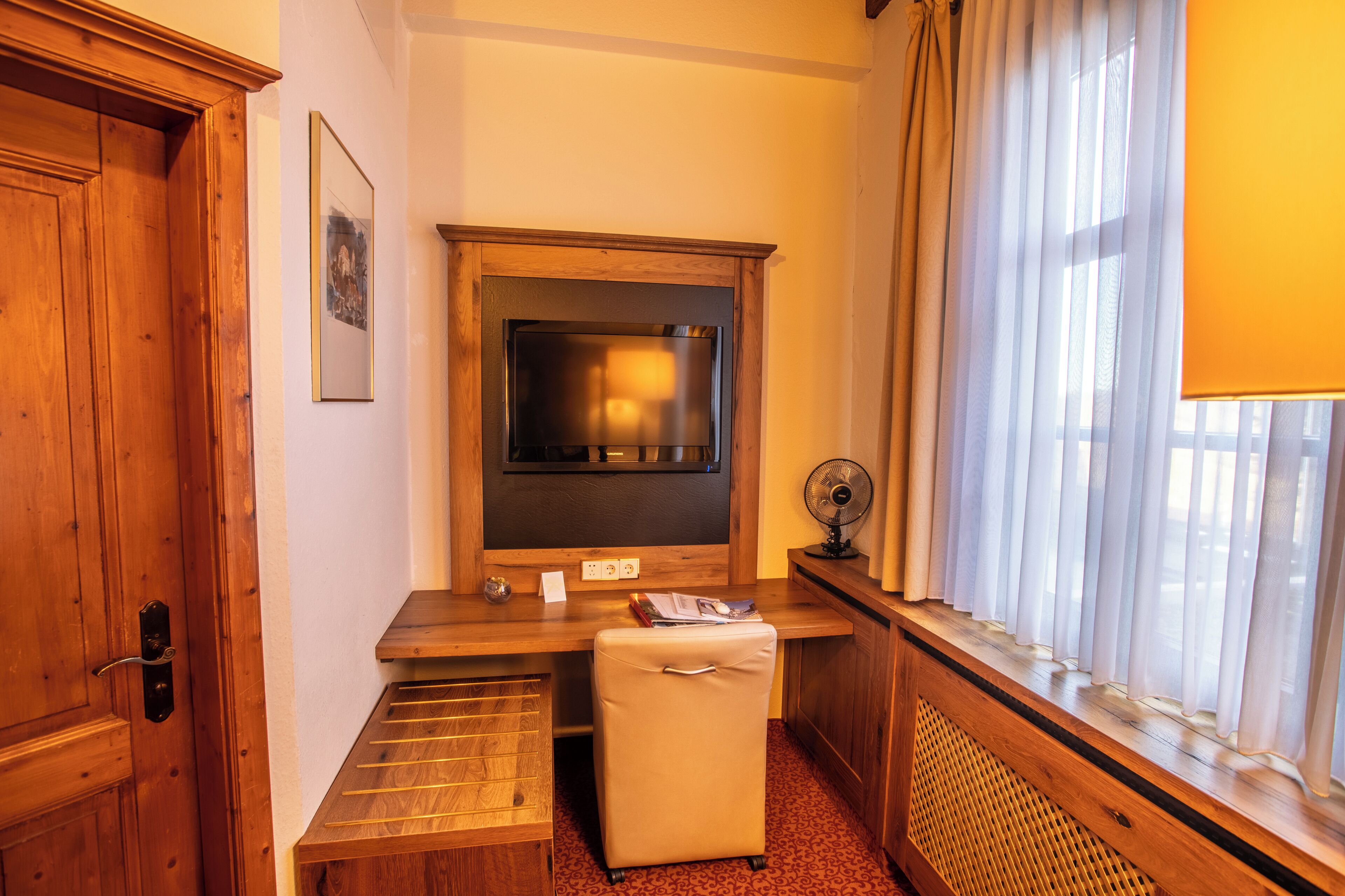 A hotel room with a wooden desk, a television, a small fan, and a window with curtains.