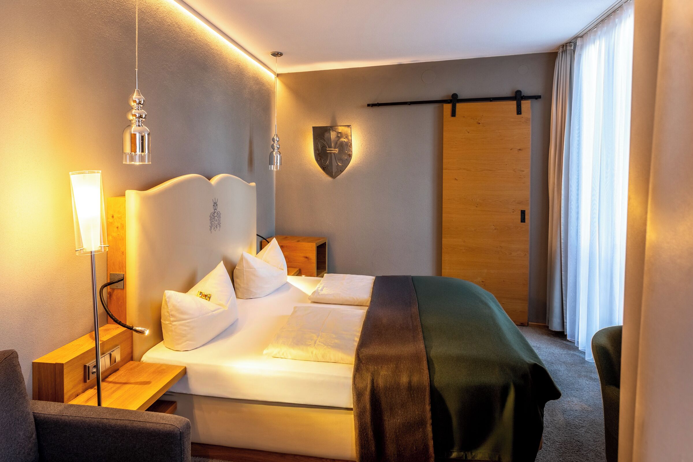 Una habitación de hotel con una cama bien hecha, una lámpara de noche, un escudo decorativo en la pared y una puerta de madera.