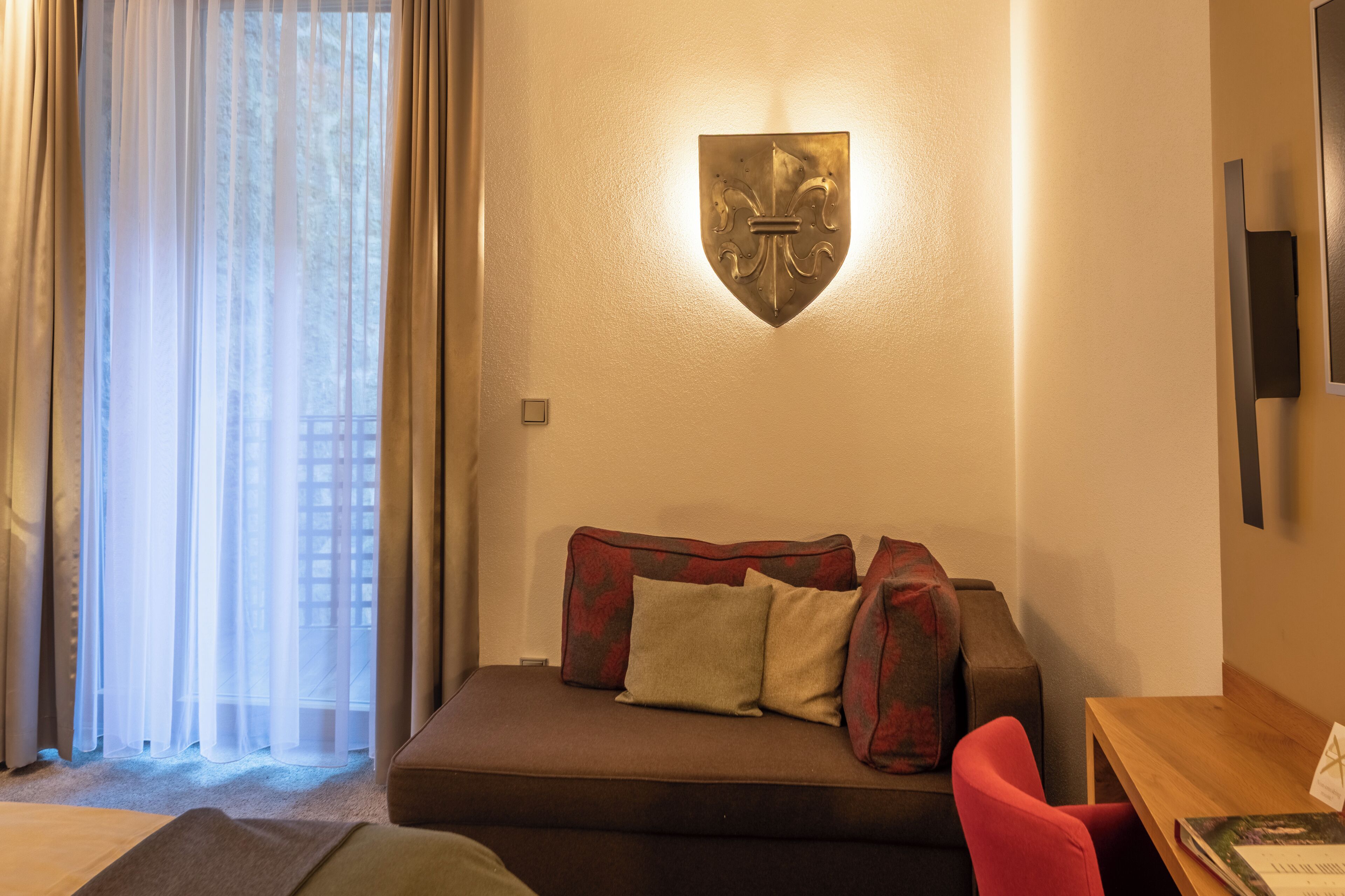 A modern hotel room with a sofa, a red chair, a wooden table, and a wall-mounted shield-shaped light fixture.