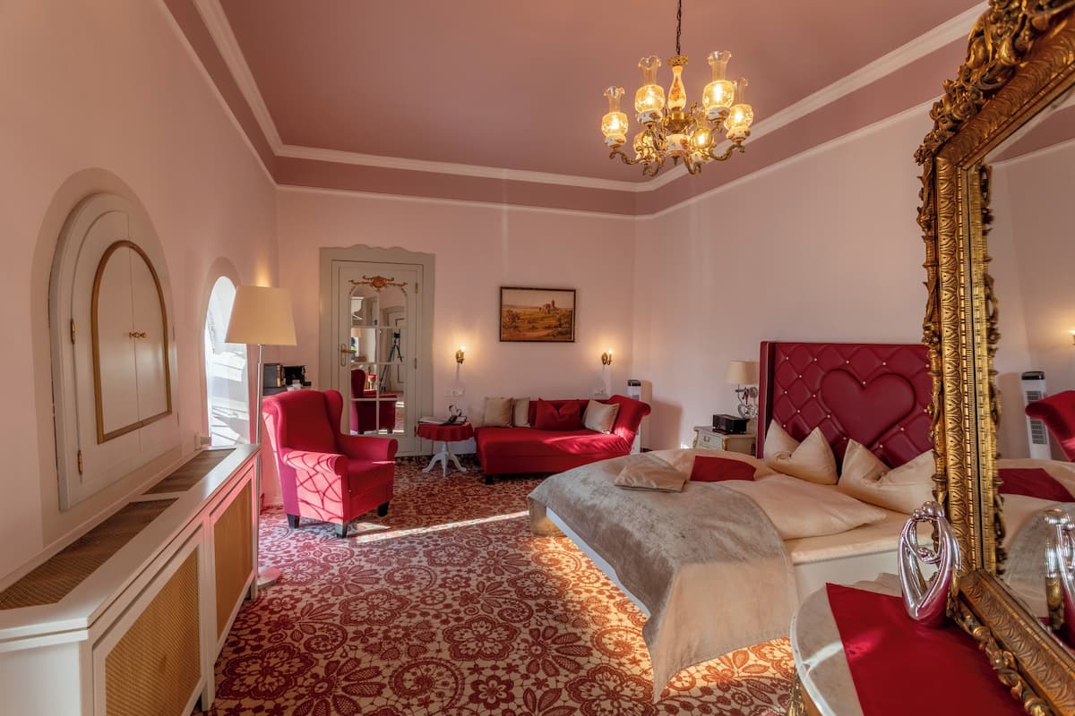 A hotel room with a red and white color scheme, a large bed, a red armchair, a small table, and a chandelier.