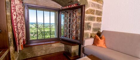 A room with a stone wall, a window with a view of greenery, a wooden bench, and a sofa with orange pillows.