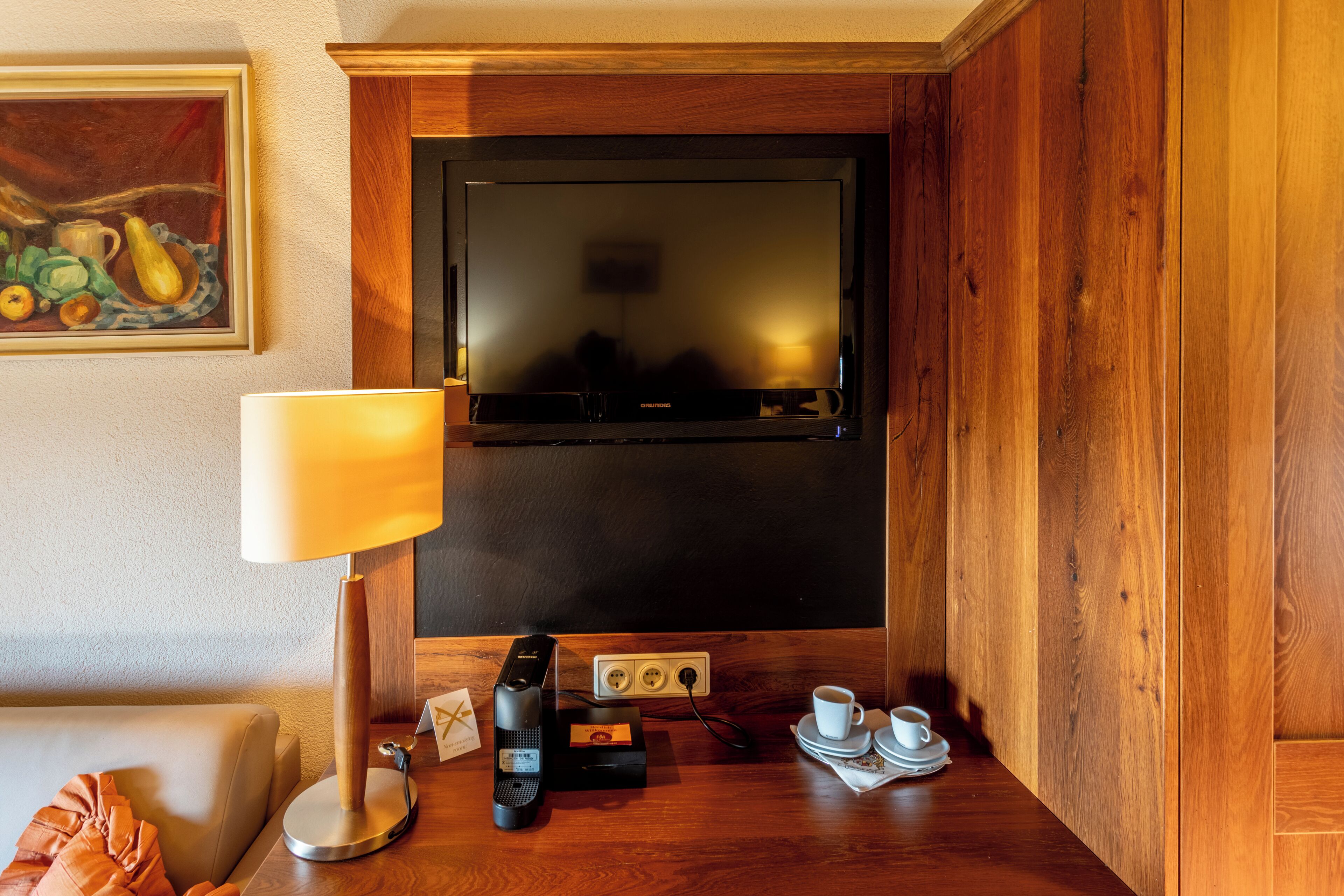 Una habitación de hotel con televisión, lámpara, cafetera y un cuadro en la pared.