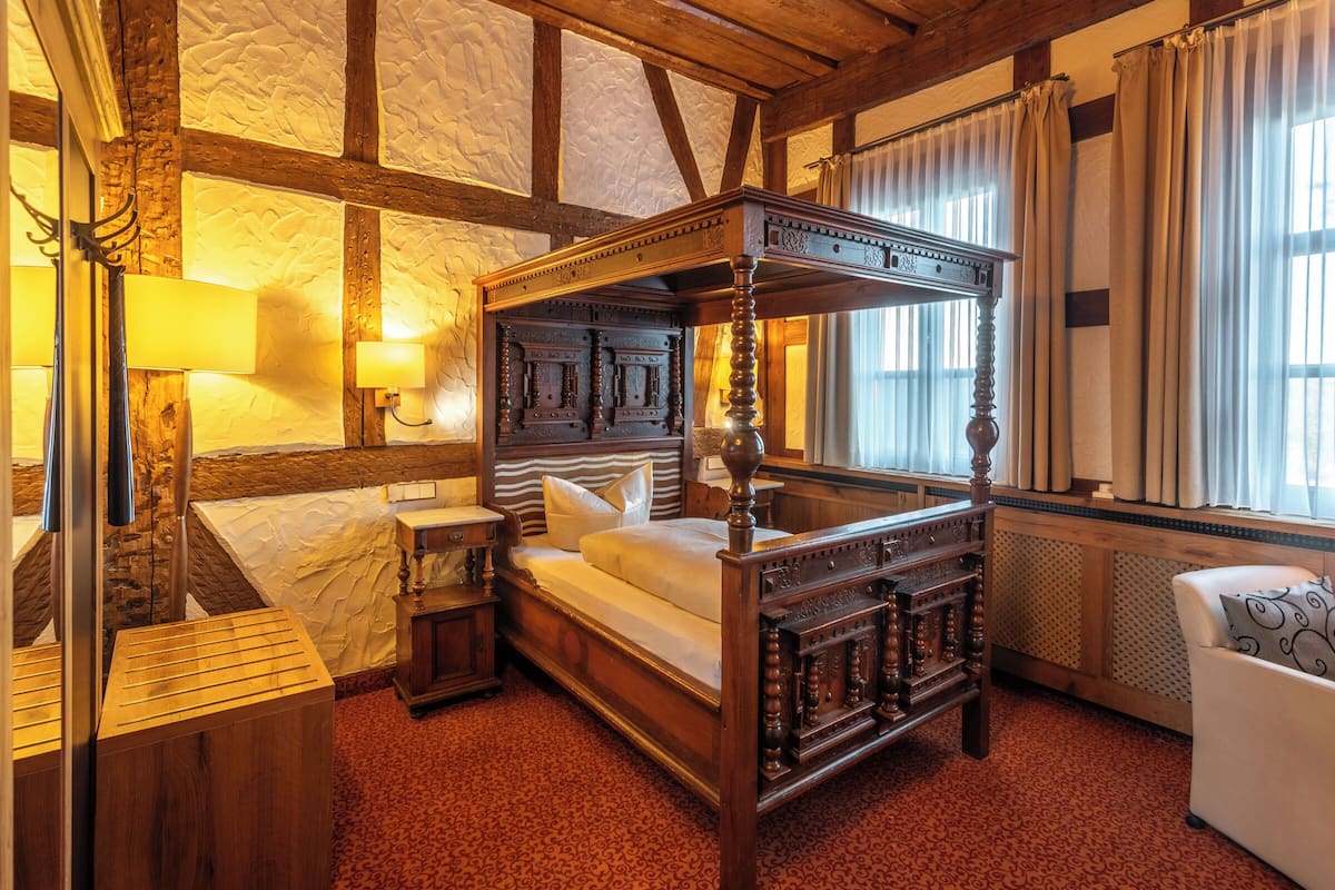 A four-poster bed with a wooden headboard, a nightstand with a lamp, a small wooden cabinet, and a white armchair.