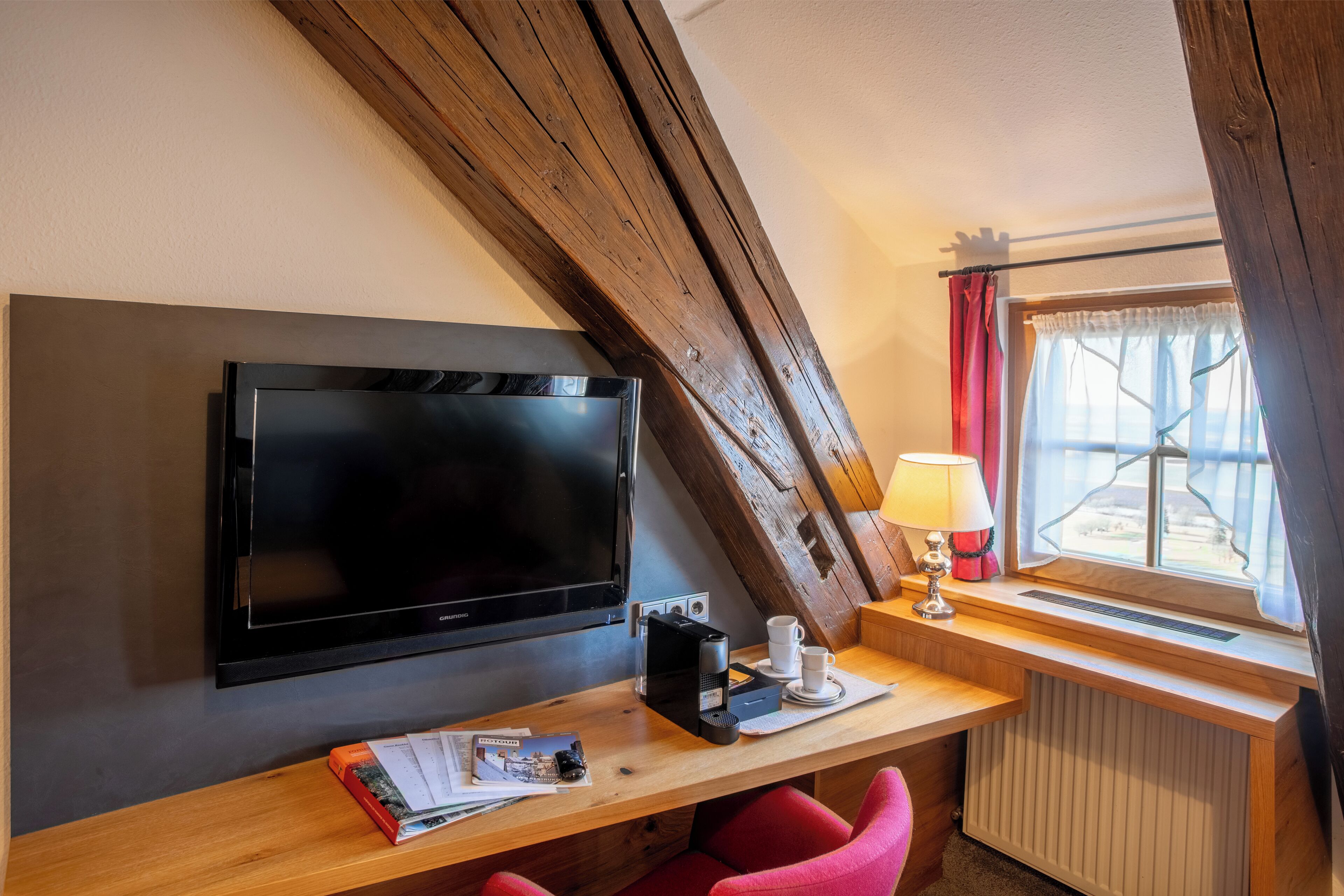 A modern room with a flat-screen TV, a wooden desk with a lamp, a red chair, and a window with curtains.
