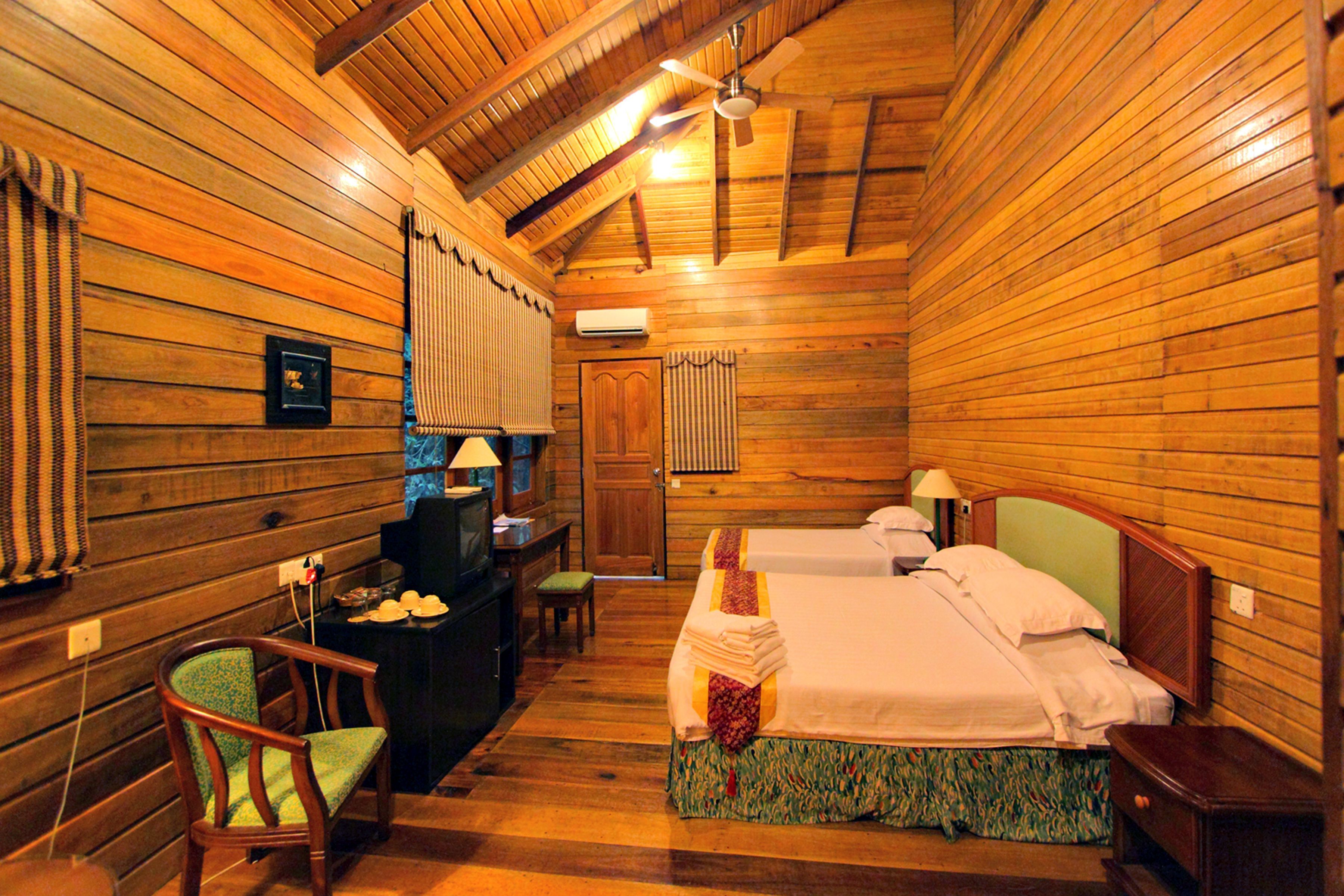 Quarto de cabana de madeira com duas camas, uma escrivaninha com luminária, uma cadeira e uma televisão.
