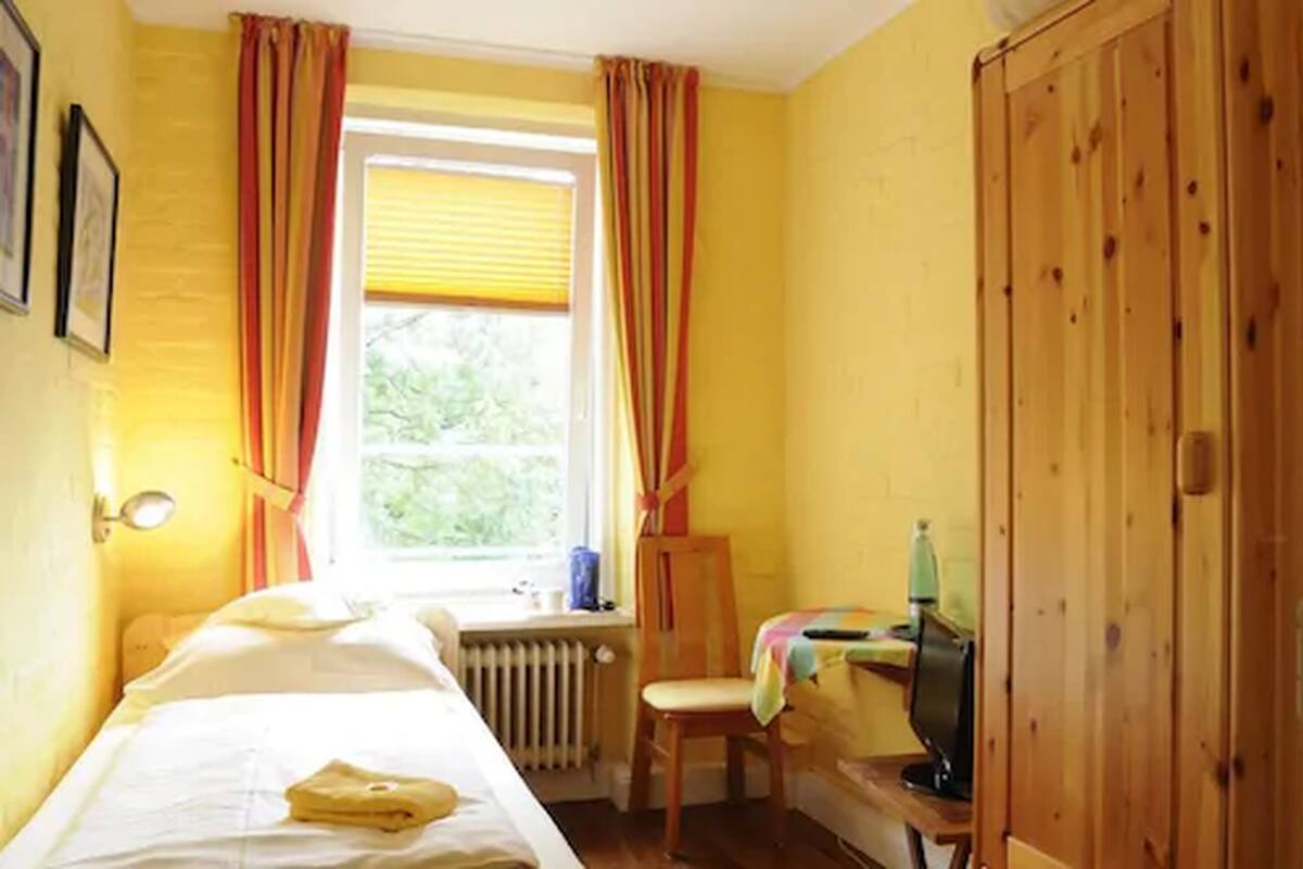 A room with a bed, a chair, a radiator, a window with curtains, and a wooden wardrobe.