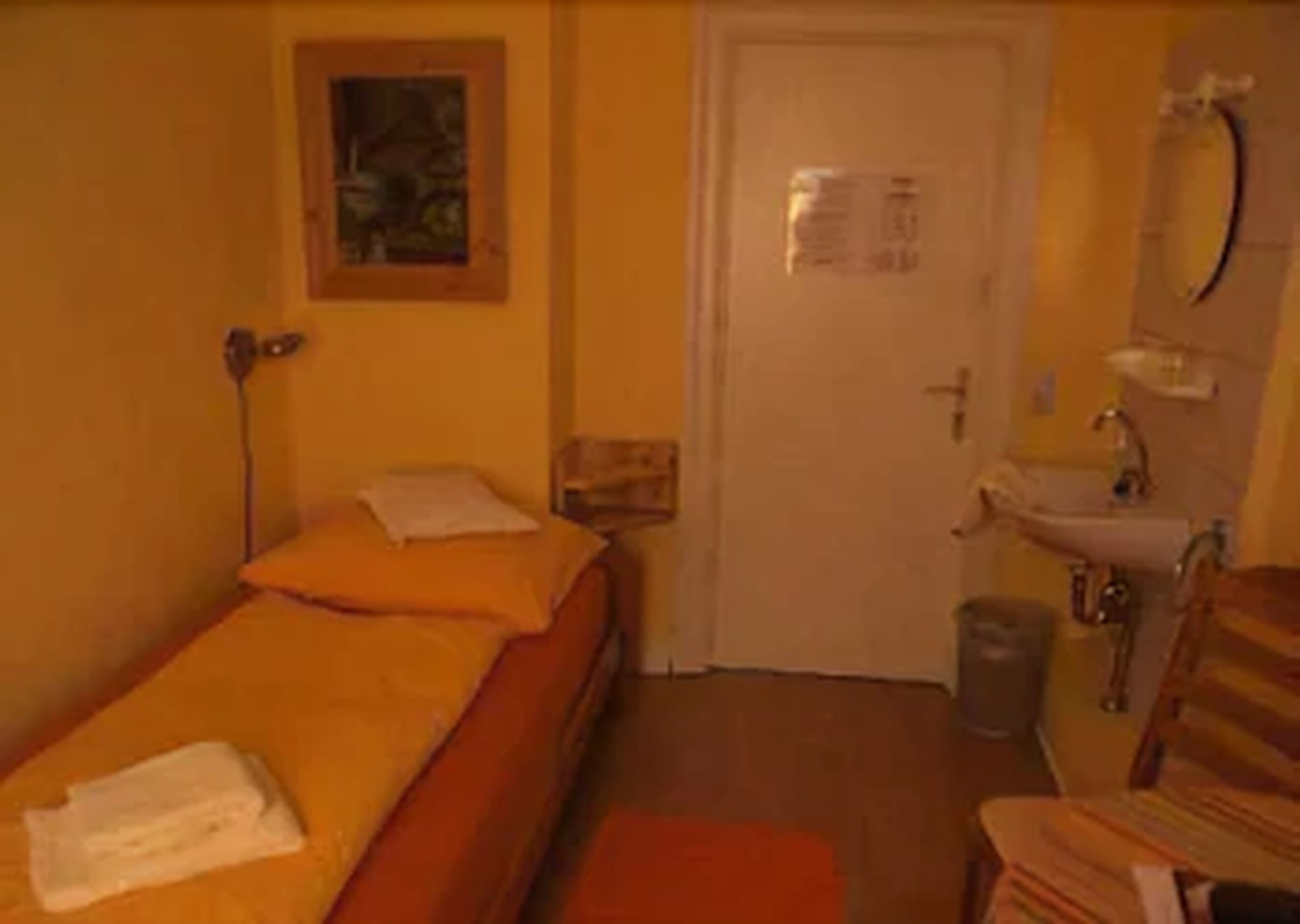 A small, single-bed room with a bed, a nightstand, a wall-mounted lamp, a door, a sink, and a chair.