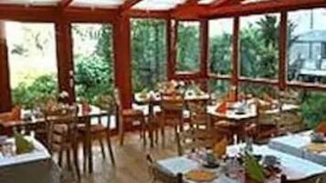 A covered outdoor dining area with tables and chairs, surrounded by large windows and greenery.