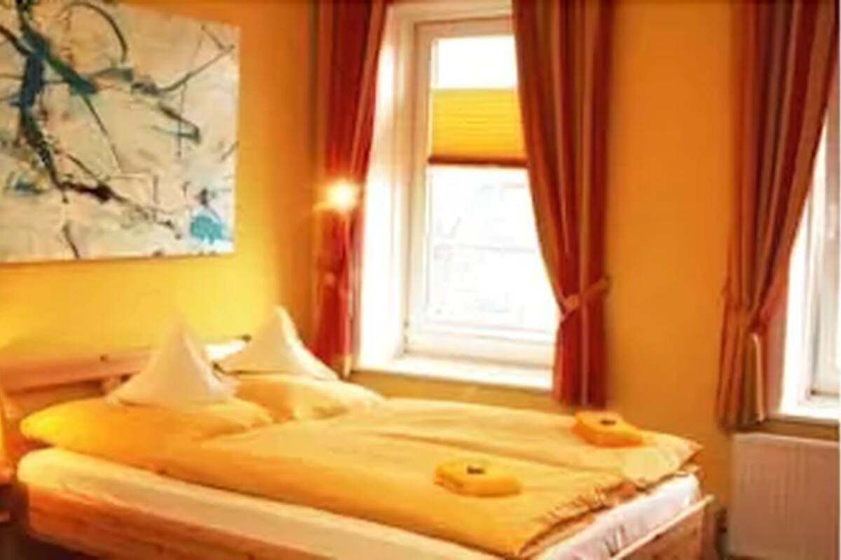 A hotel room with a bed, pillows, a window with curtains, and a painting on the wall.