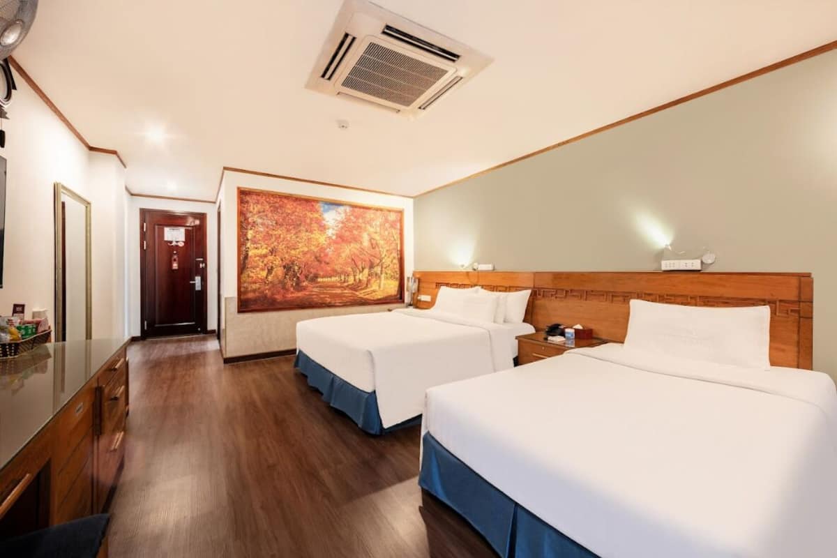 Une chambre d’hôtel avec deux lits, une tête de lit en bois, un grand tableau représentant des feuilles d’automne, un bureau avec un miroir et un appareil de climatisation.