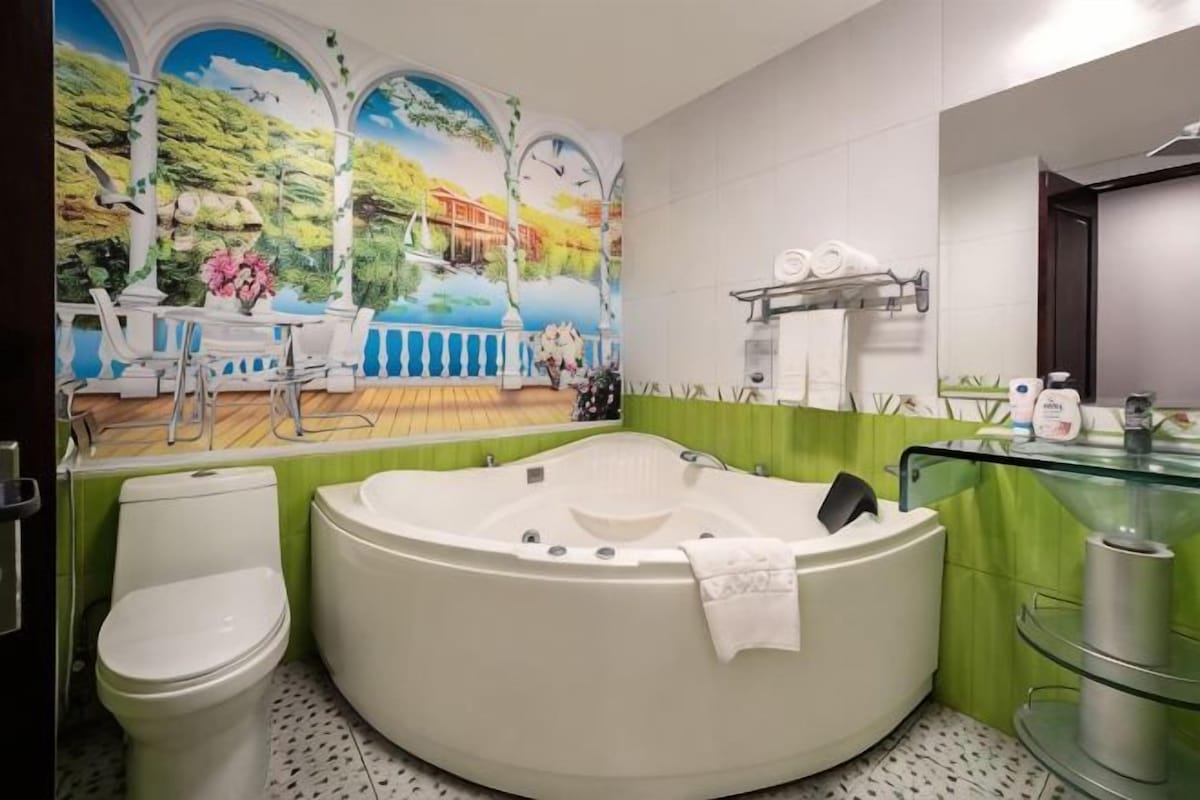 Um banheiro com uma banheira de hidromassagem grande, vaso sanitário, pia de vidro e um mural com vista para um lago.