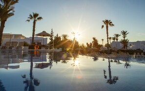 A pool with sun reflections, palm trees, lounge chairs, and a building with a sign.