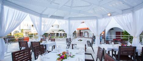 An outdoor dining area with white tables and chairs, covered by a white canopy.