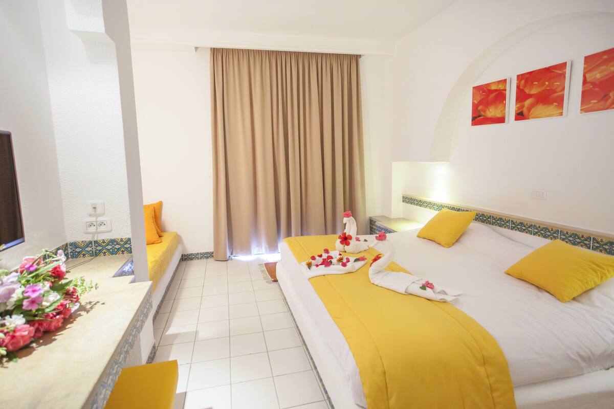 A hotel room with a bed, a desk, and a chair. There is a window with curtains, a wall with artwork, and a tiled floor.