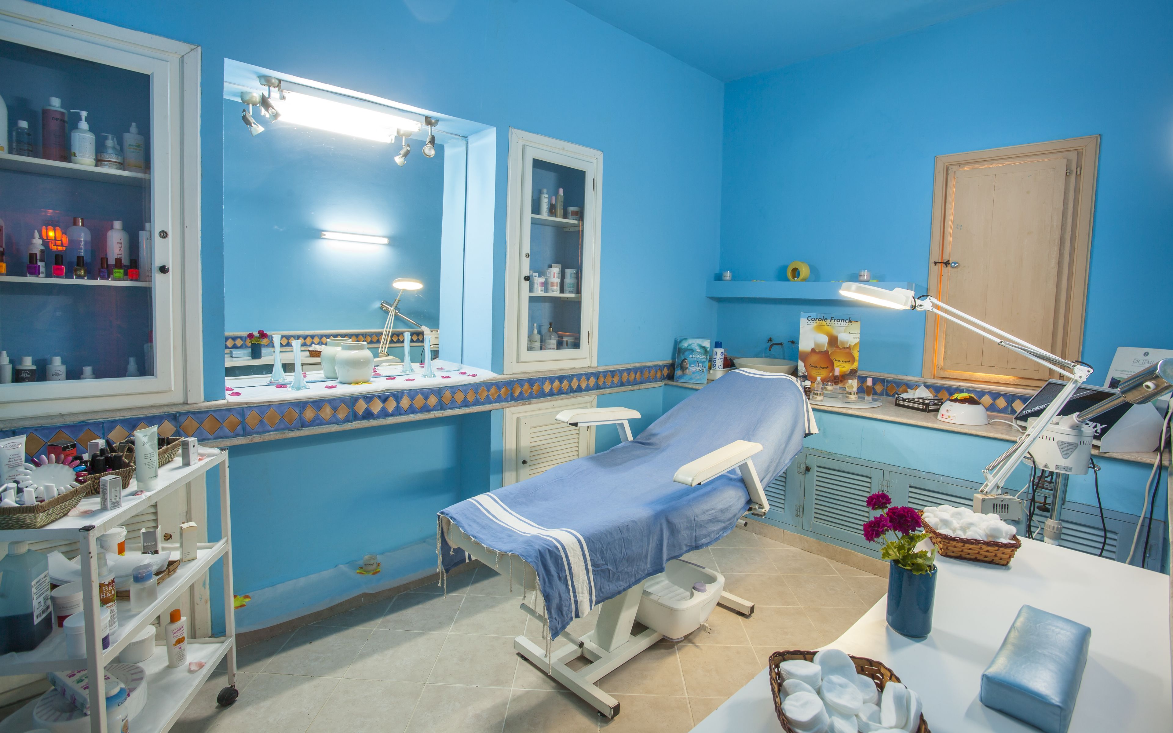 A medical examination room with a bed, examination table, shelves with medical supplies, a cabinet, and a desk with a computer and lamp.