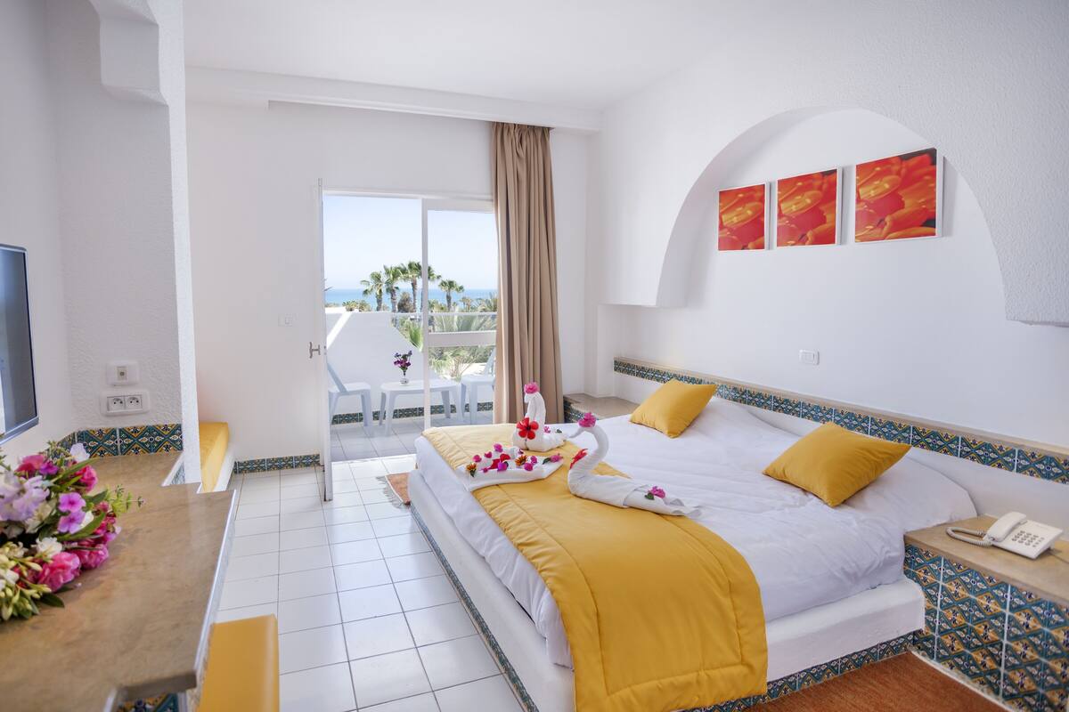A hotel room with a bed, a desk, a chair, a TV, and a balcony with a view of the sea.