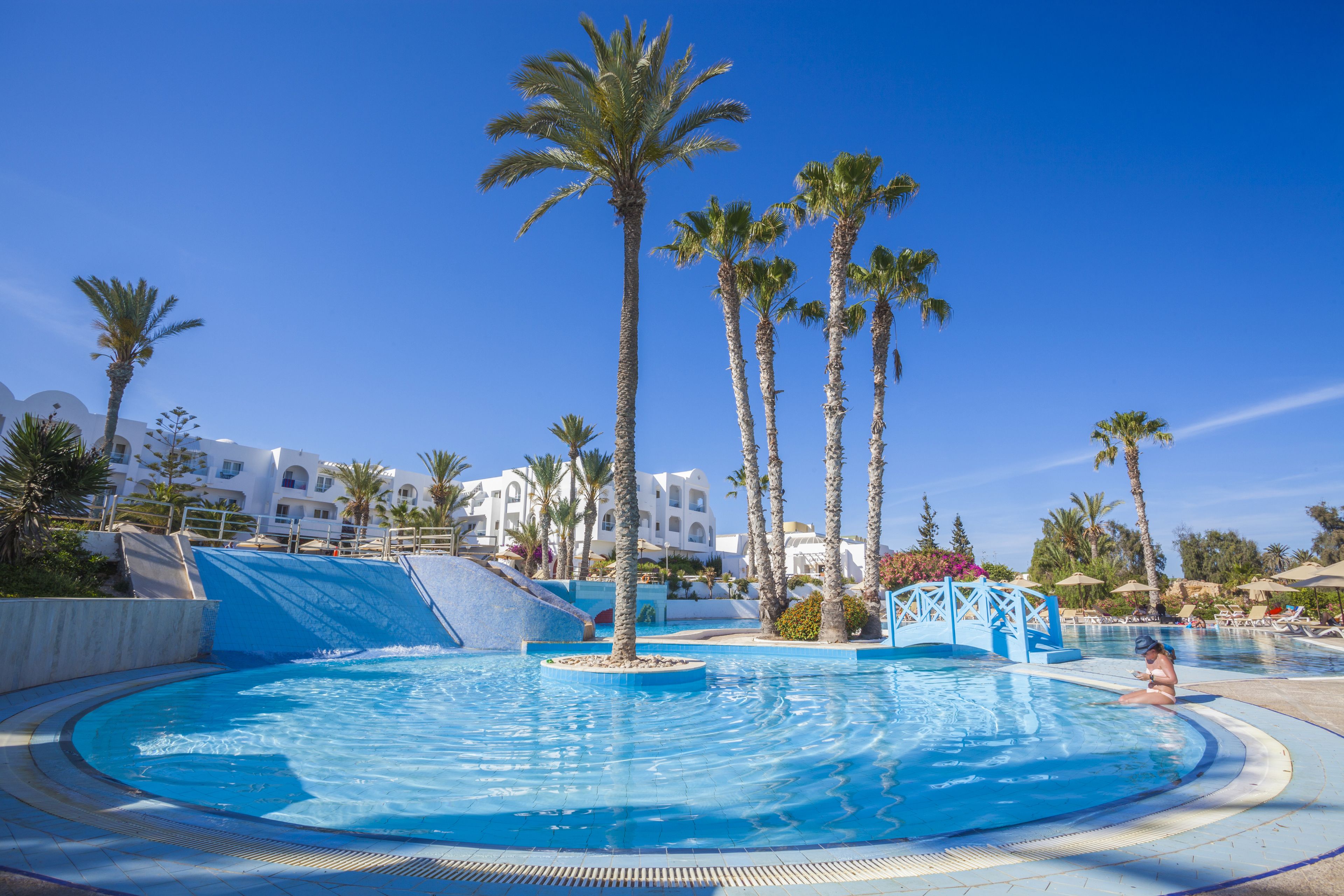 Une piscine bordée de palmiers, un toboggan aquatique et, au fond, un bâtiment doté de balcons.